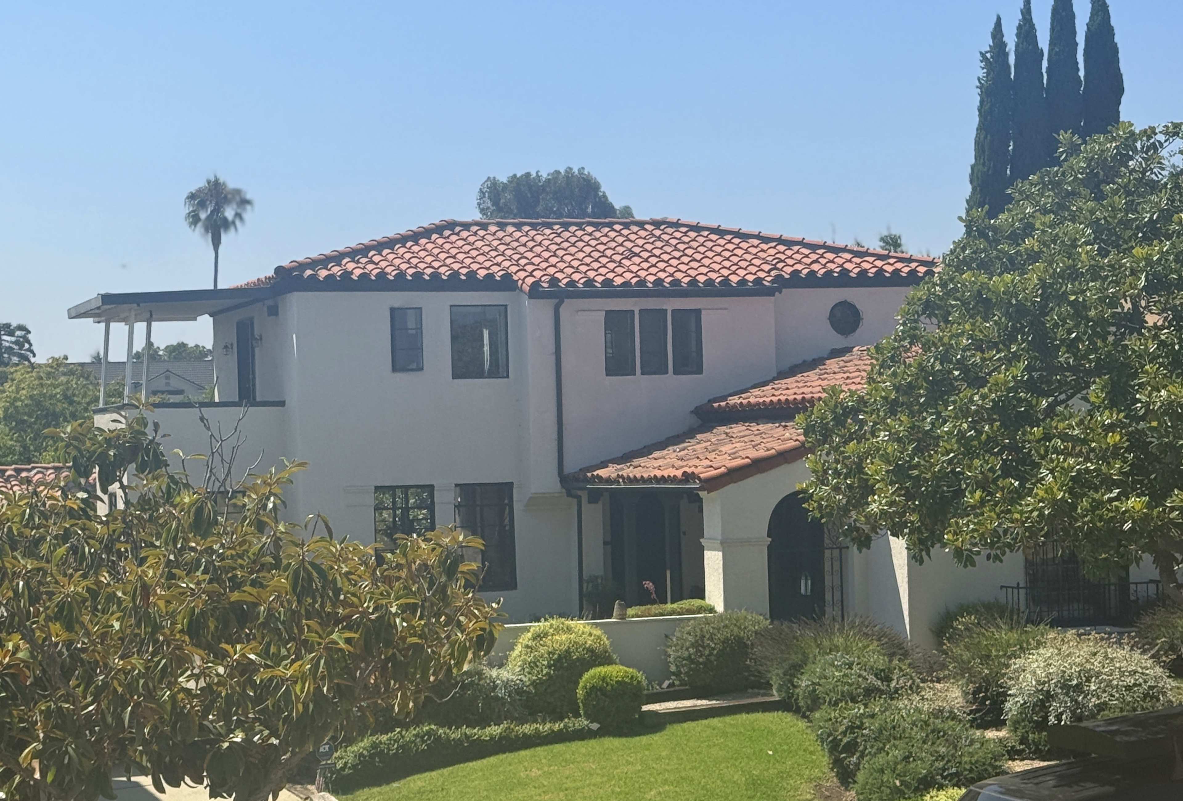 A two-story house features a red tile roof, white stucco exterior, and landscaped grounds with various shrubs and trees.
