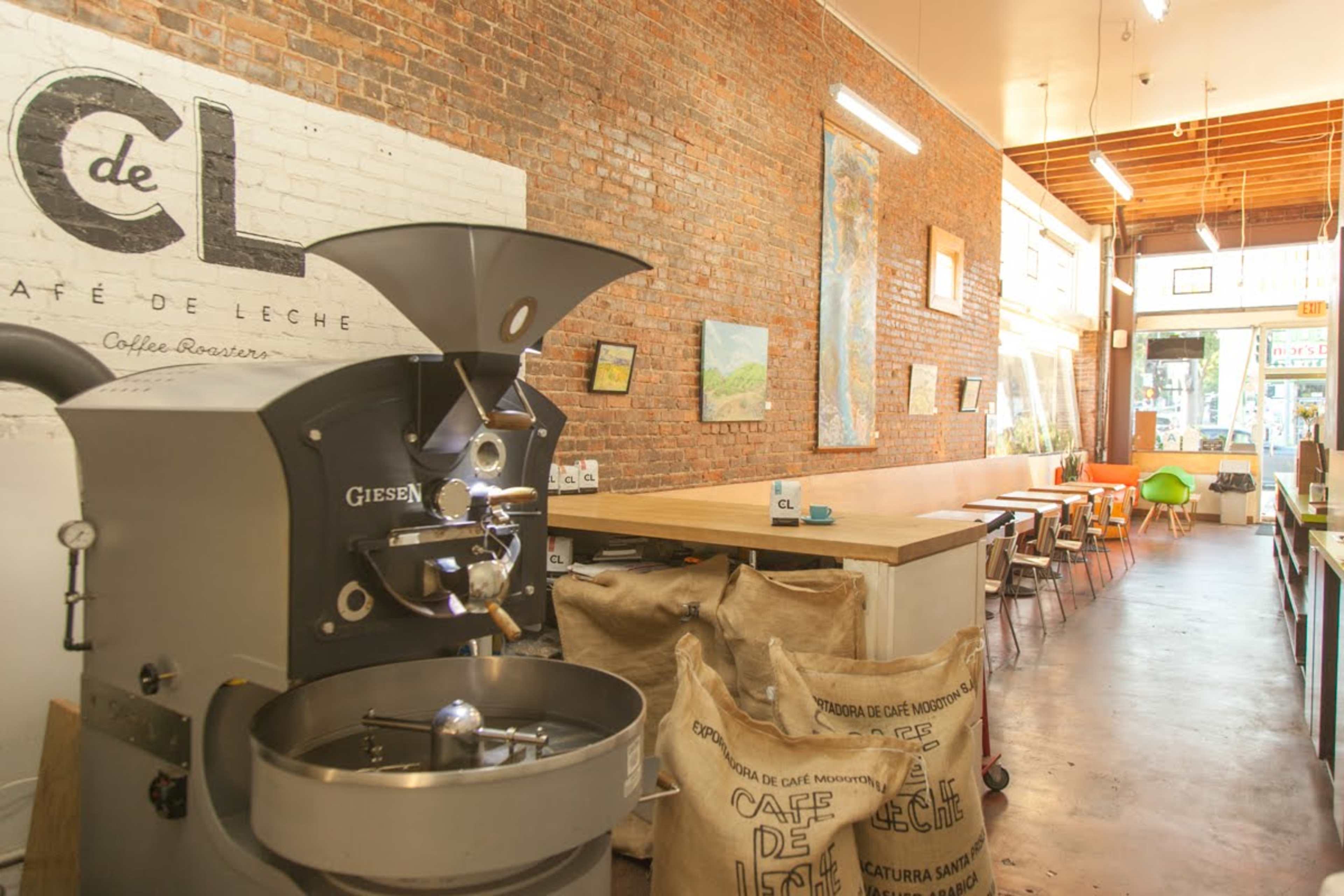 A coffee roasting shop with a Giesen coffee roaster, wooden communal tables, and a brick wall adorned with art.