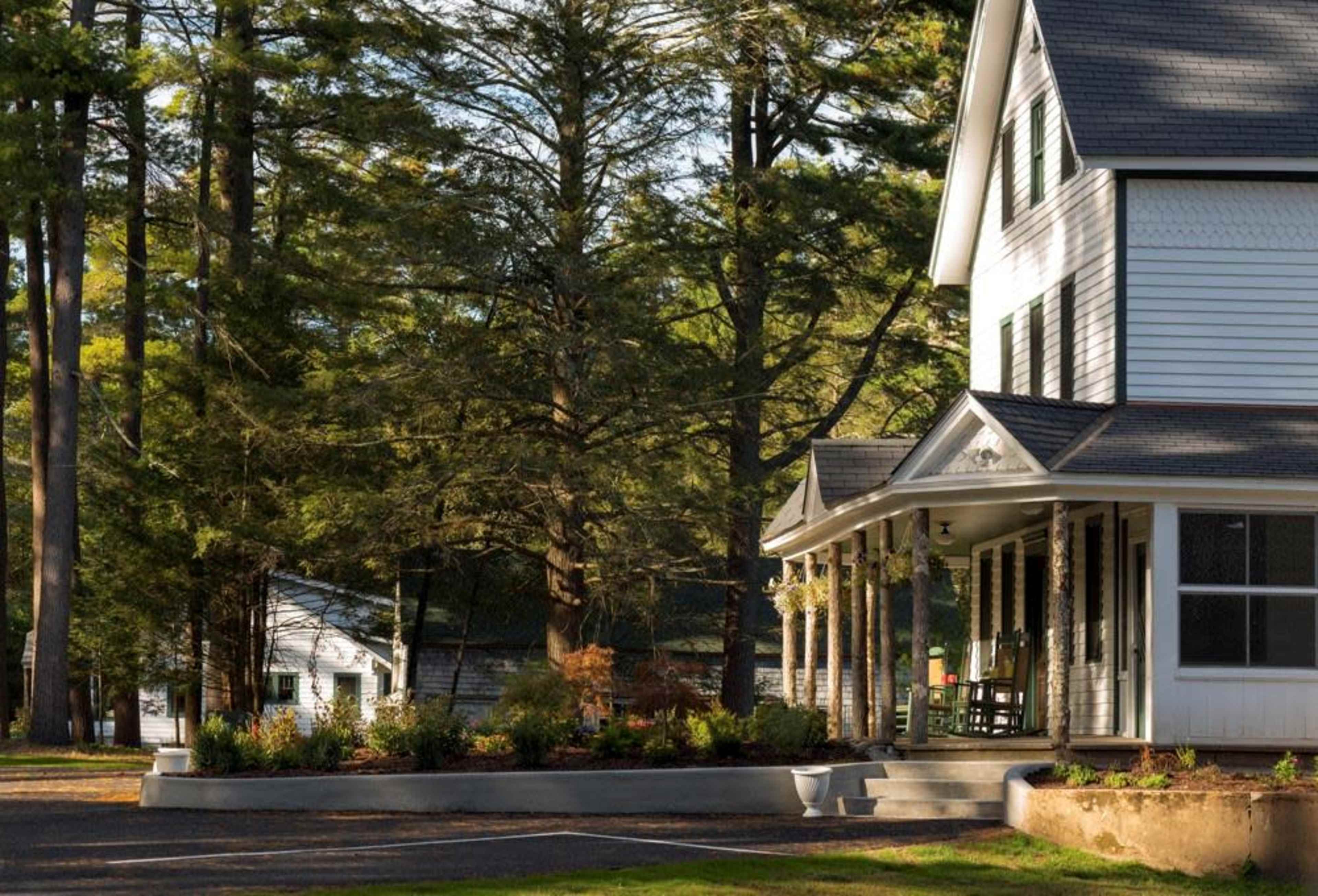 A two-story white house with a porch and a view of tall trees is situated alongside a smaller building in a wooded area.