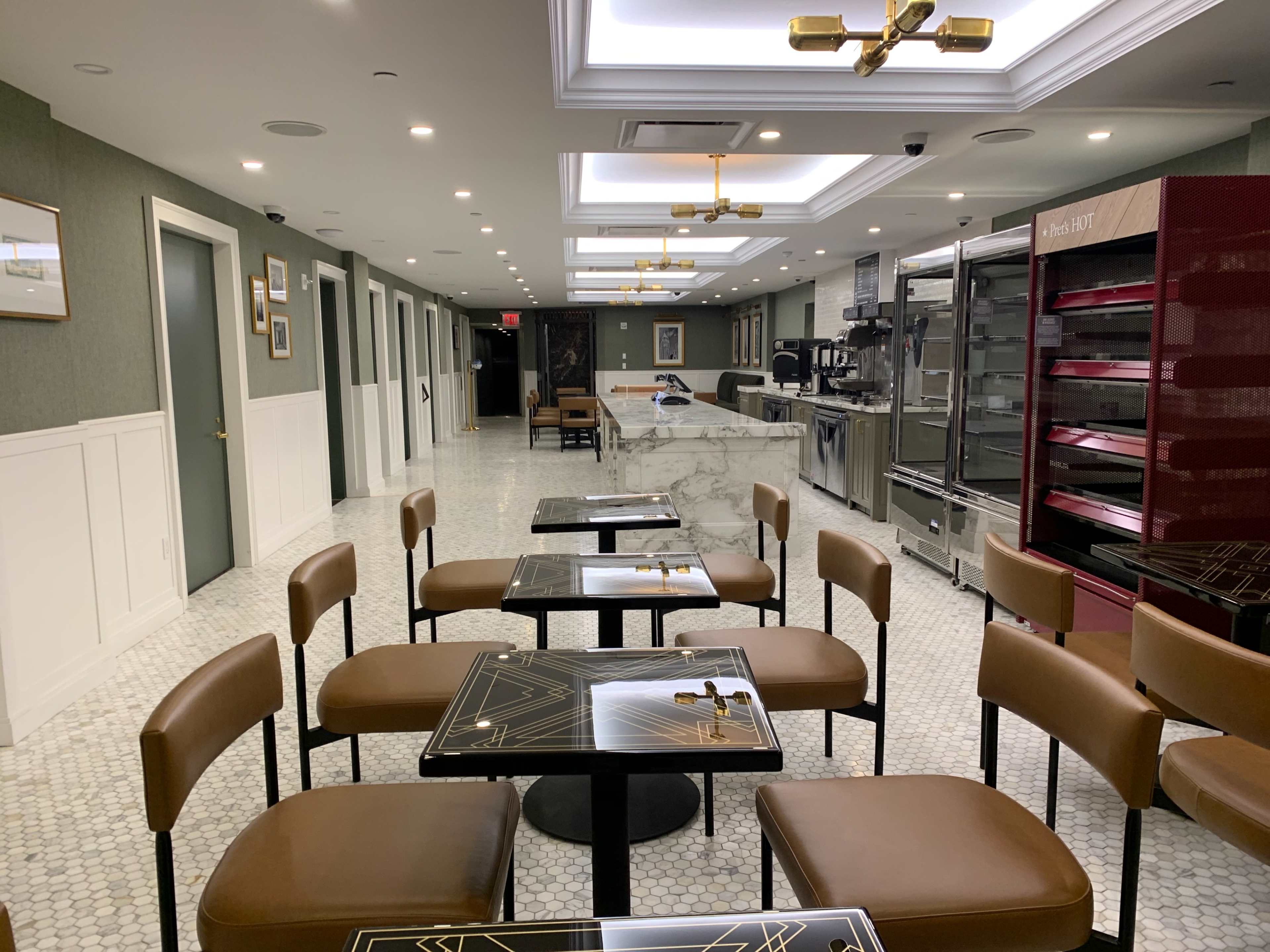 The image shows an empty modern café with brown chairs and tables, featuring a marble counter and a display area in the background.