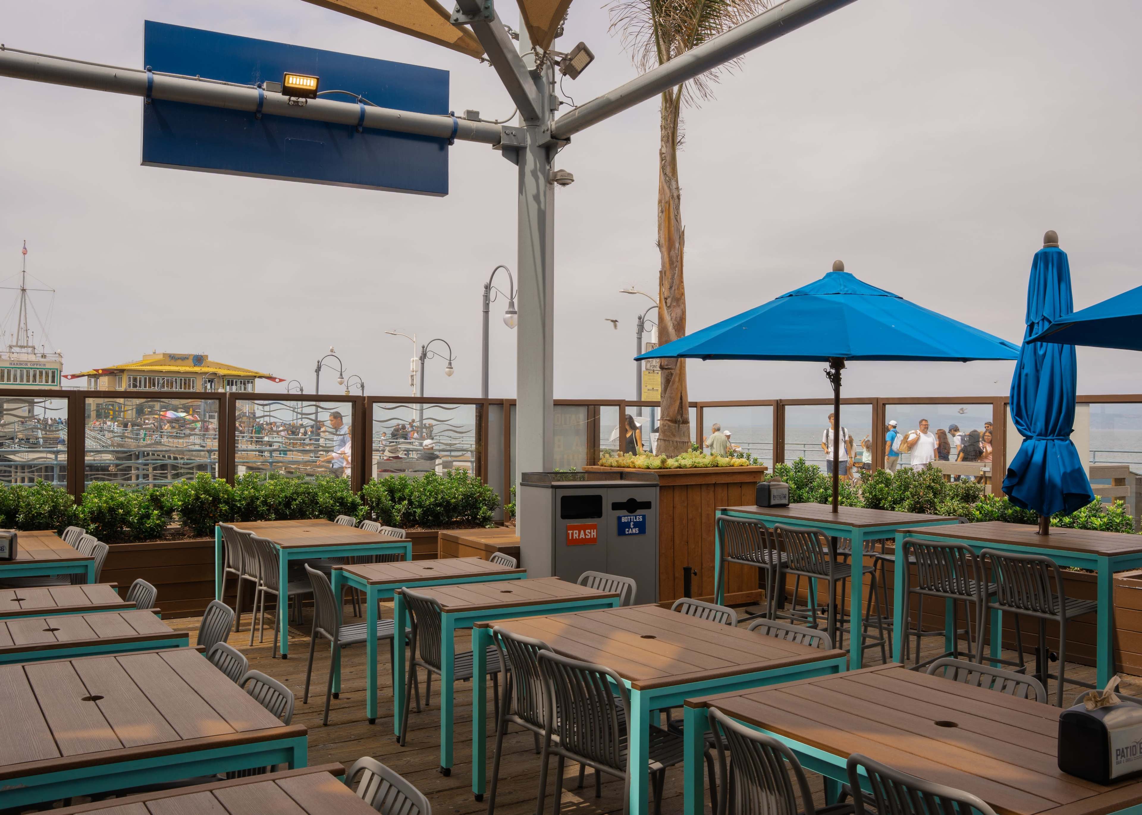 The image shows an outdoor dining area with blue umbrellas and wooden tables overlooking a harbor with boats in the background.