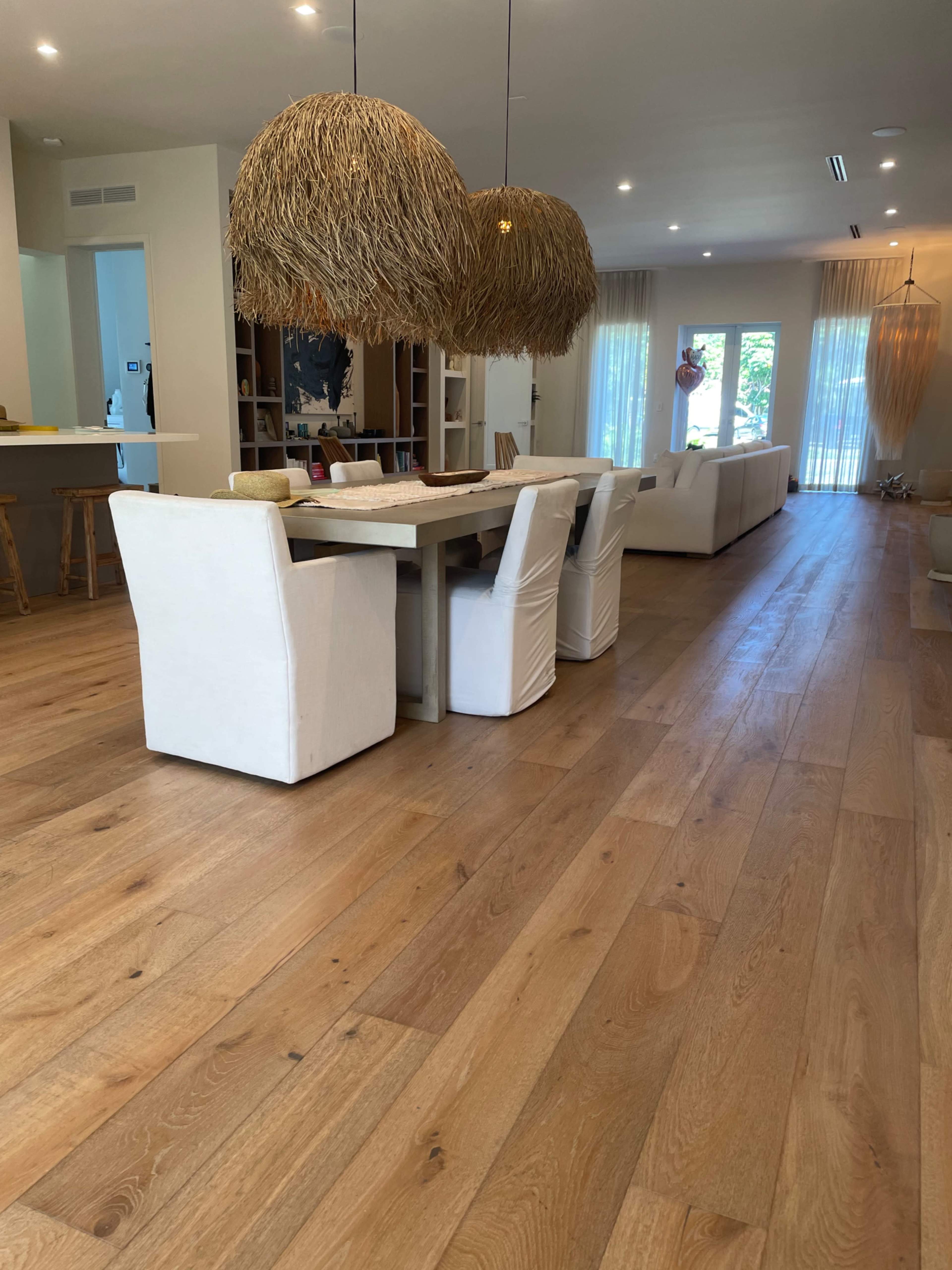 The image shows a spacious modern living area with a long dining table, upholstered chairs, and large pendant lights hanging overhead.