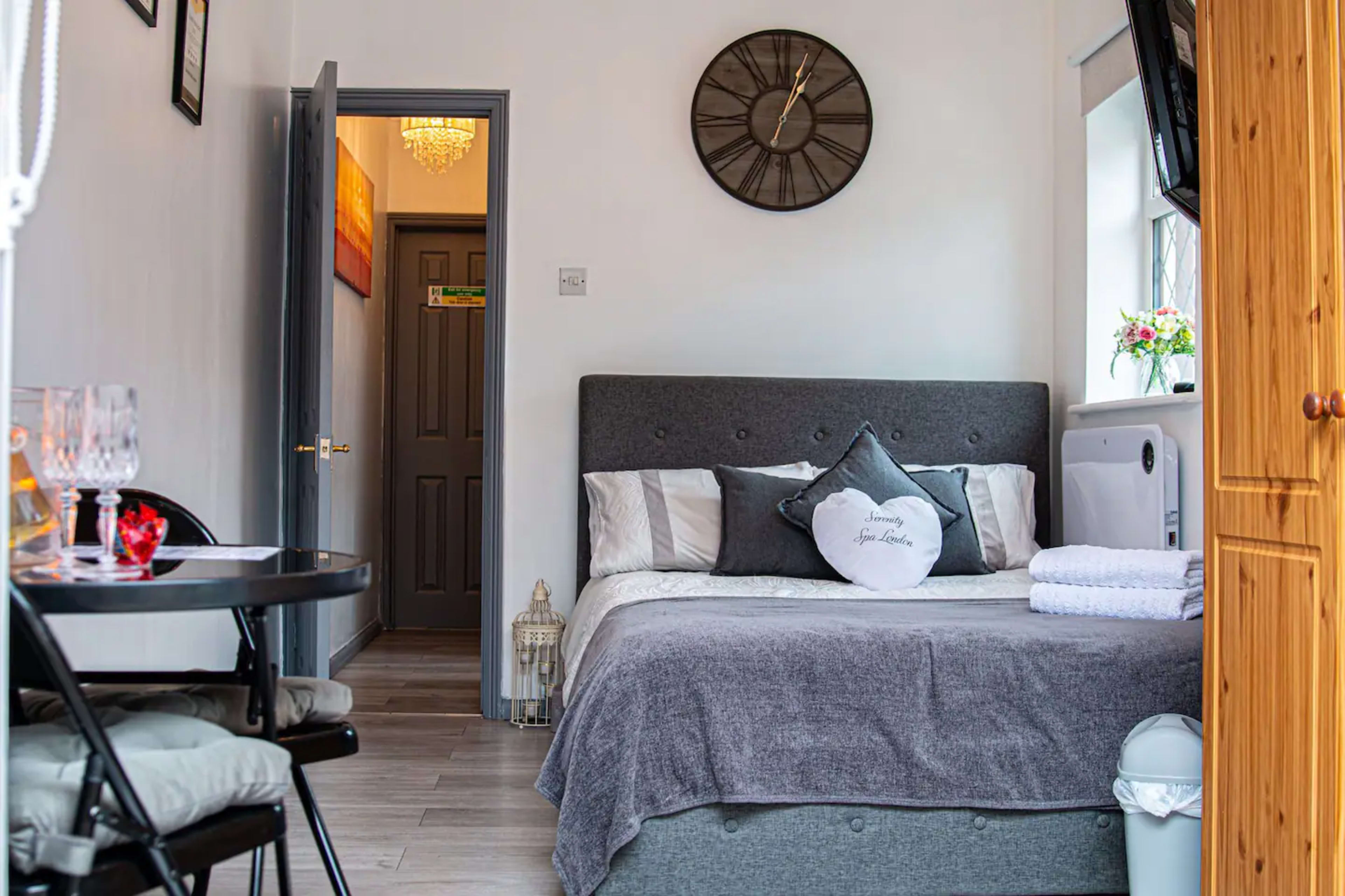 A compact room featuring a bed with gray bedding, a clock on the wall, a small dining table with chairs, and a doorway leading to another room.