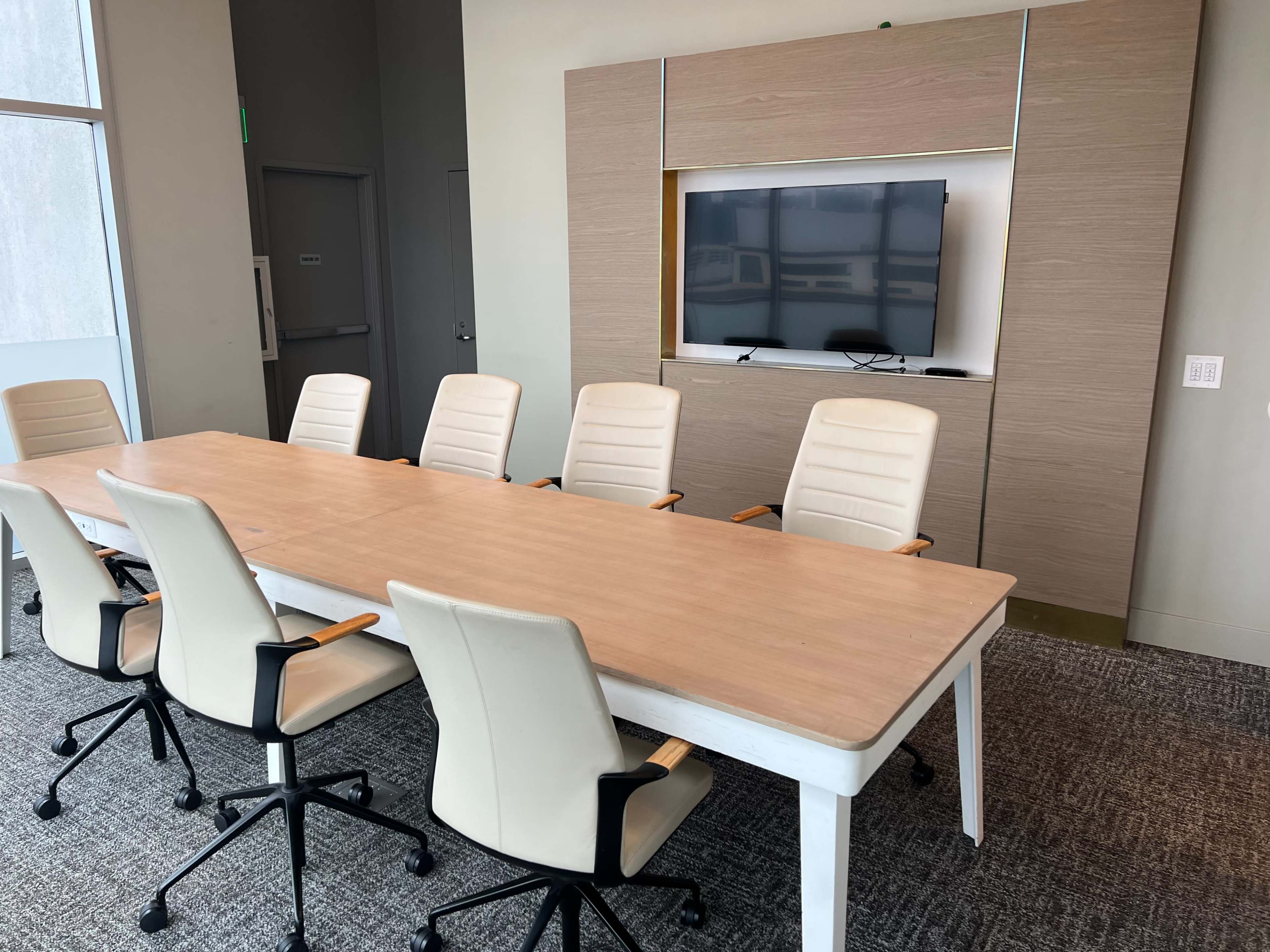 A conference room features a long wooden table surrounded by six white office chairs, with a large television mounted on a wall unit behind it.