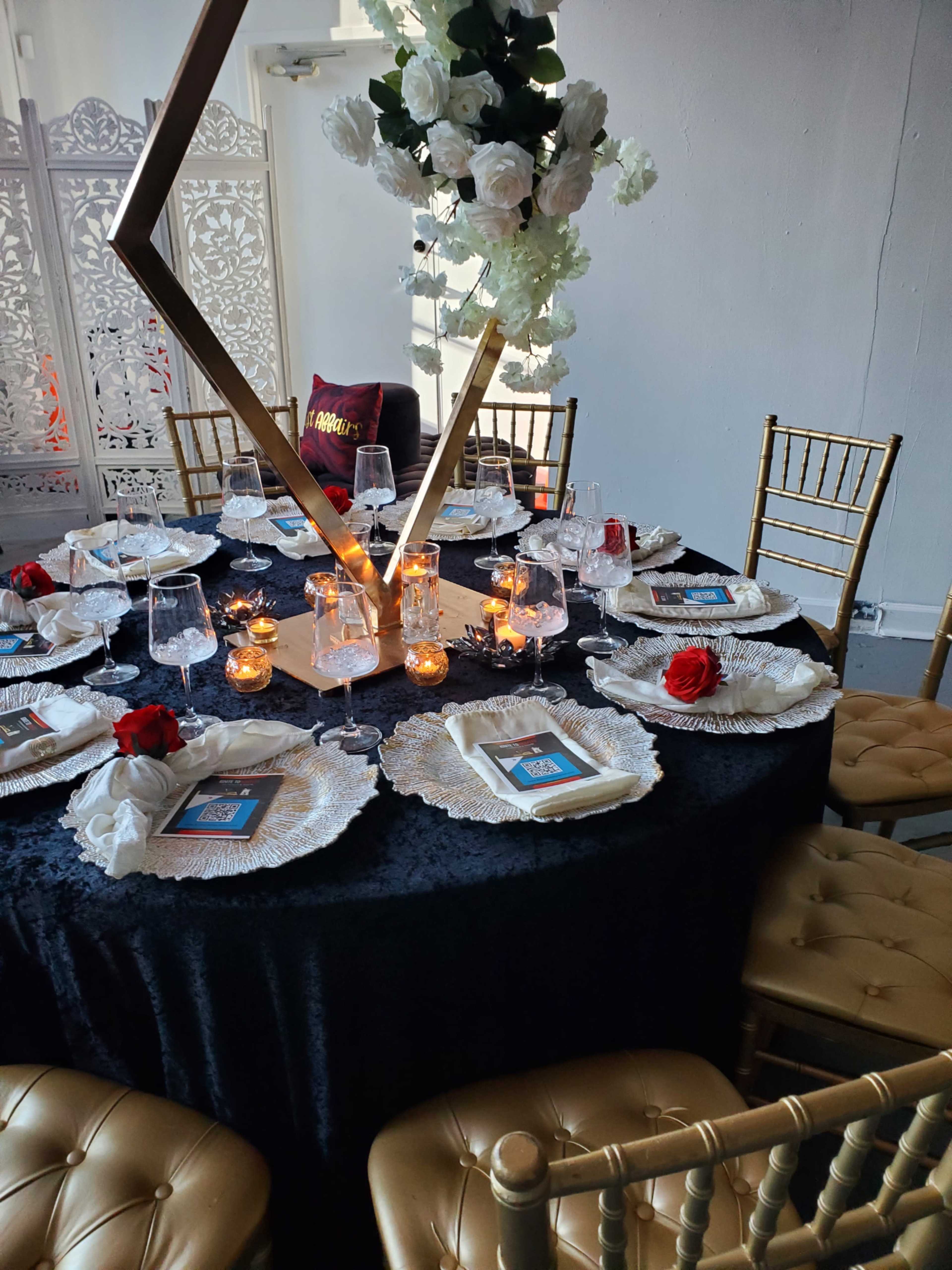 A round dining table is elegantly set with black and gold décor, featuring white plates, glassware, and centerpieces of candles and flowers.