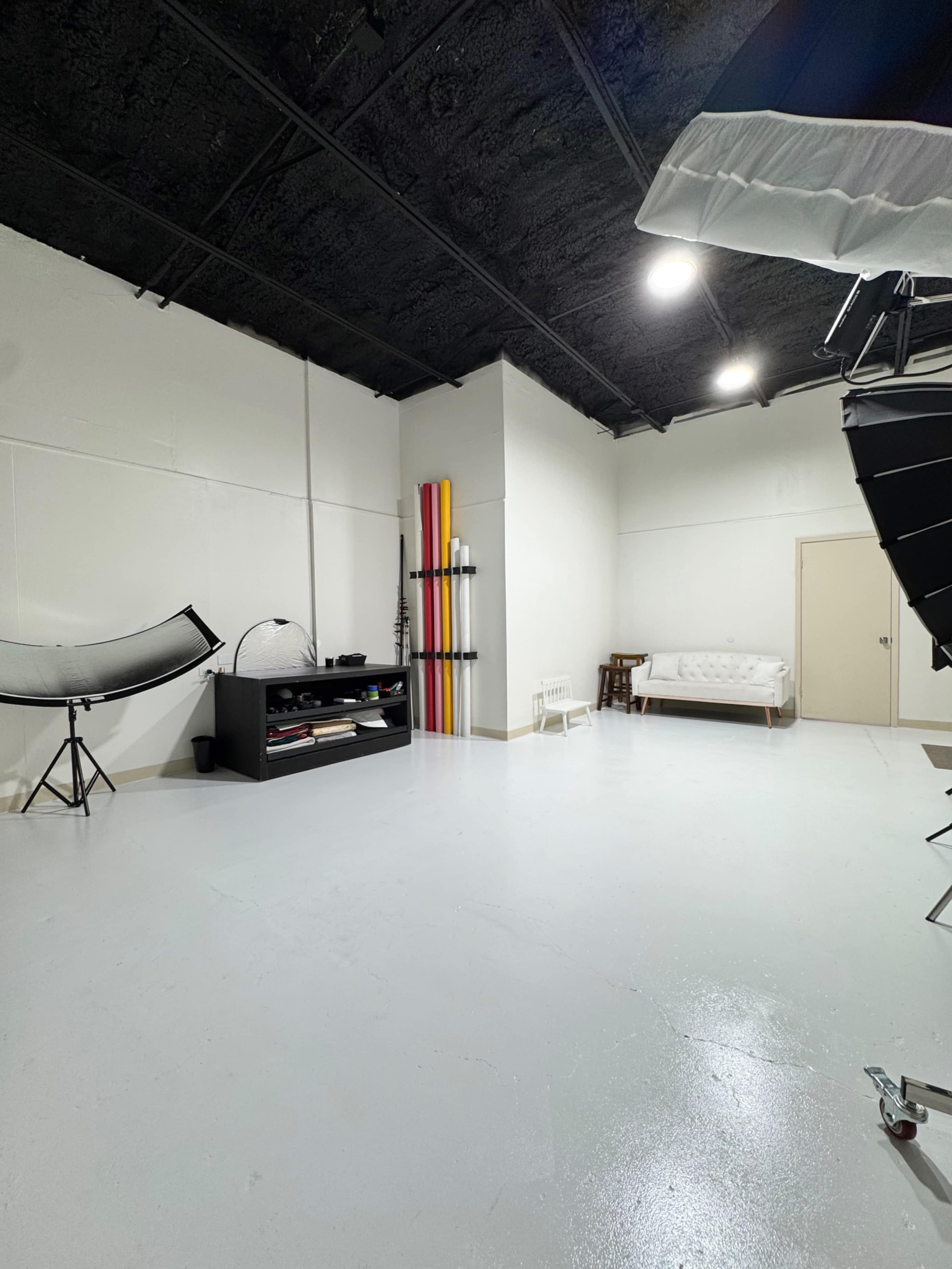The image shows an empty photography studio with a white floor, a sofa against the wall, and various photo equipment arranged around the space.