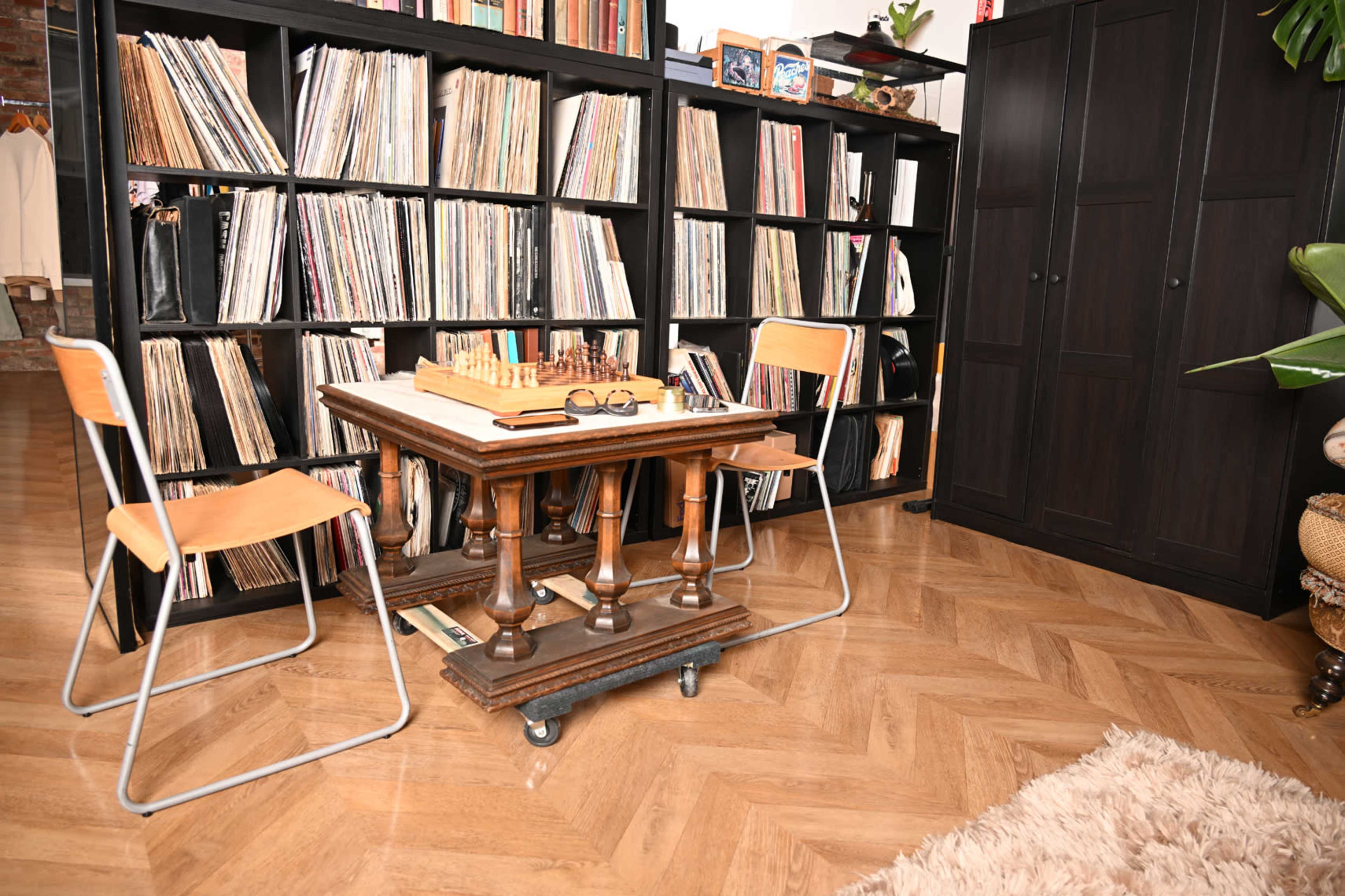 The image features a wooden chess table with two chairs, surrounded by tall black bookshelves filled with vinyl records.