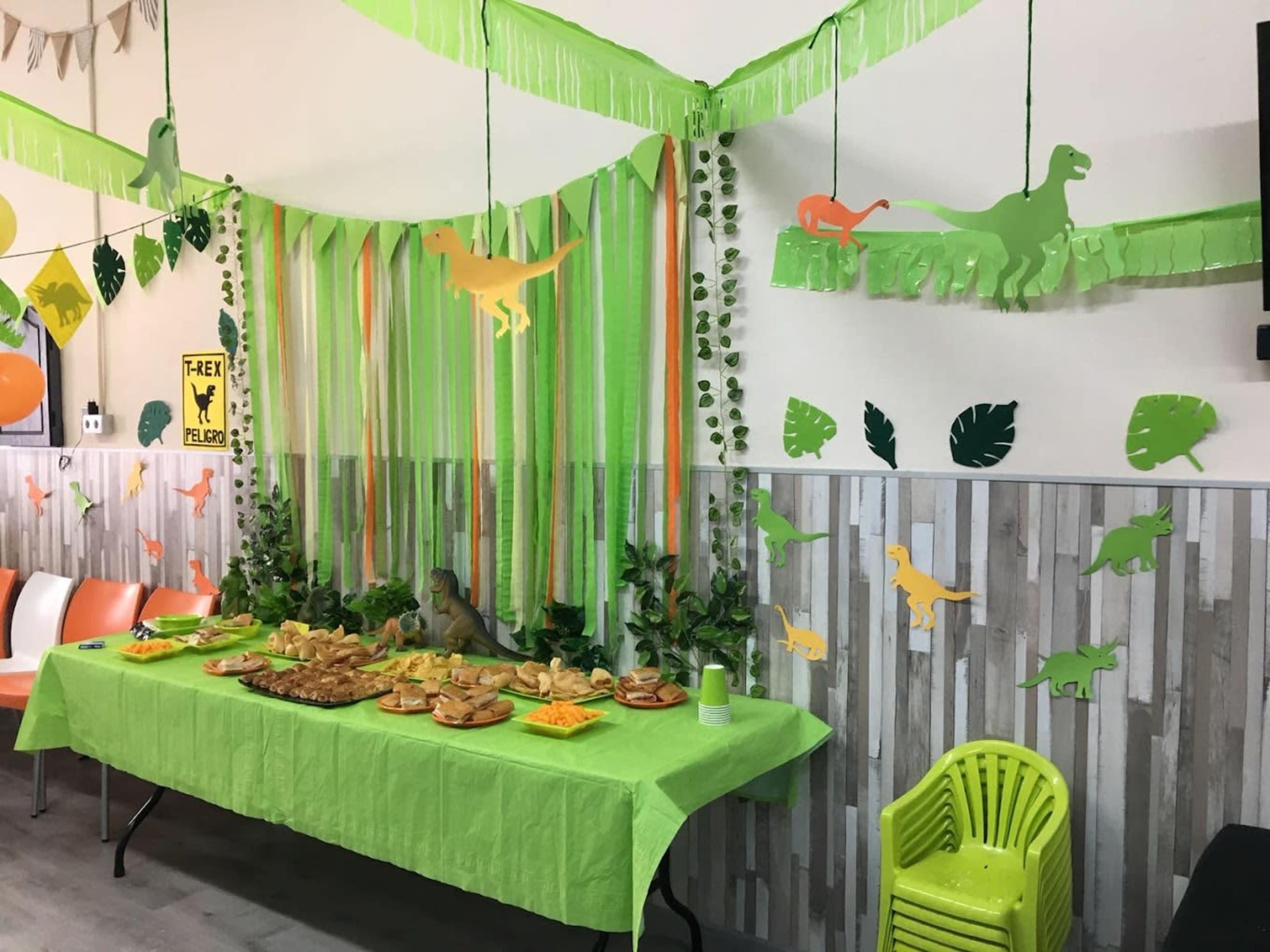 A themed party area features a green tablecloth with a spread of food, surrounded by dinosaur decorations and green streamers.