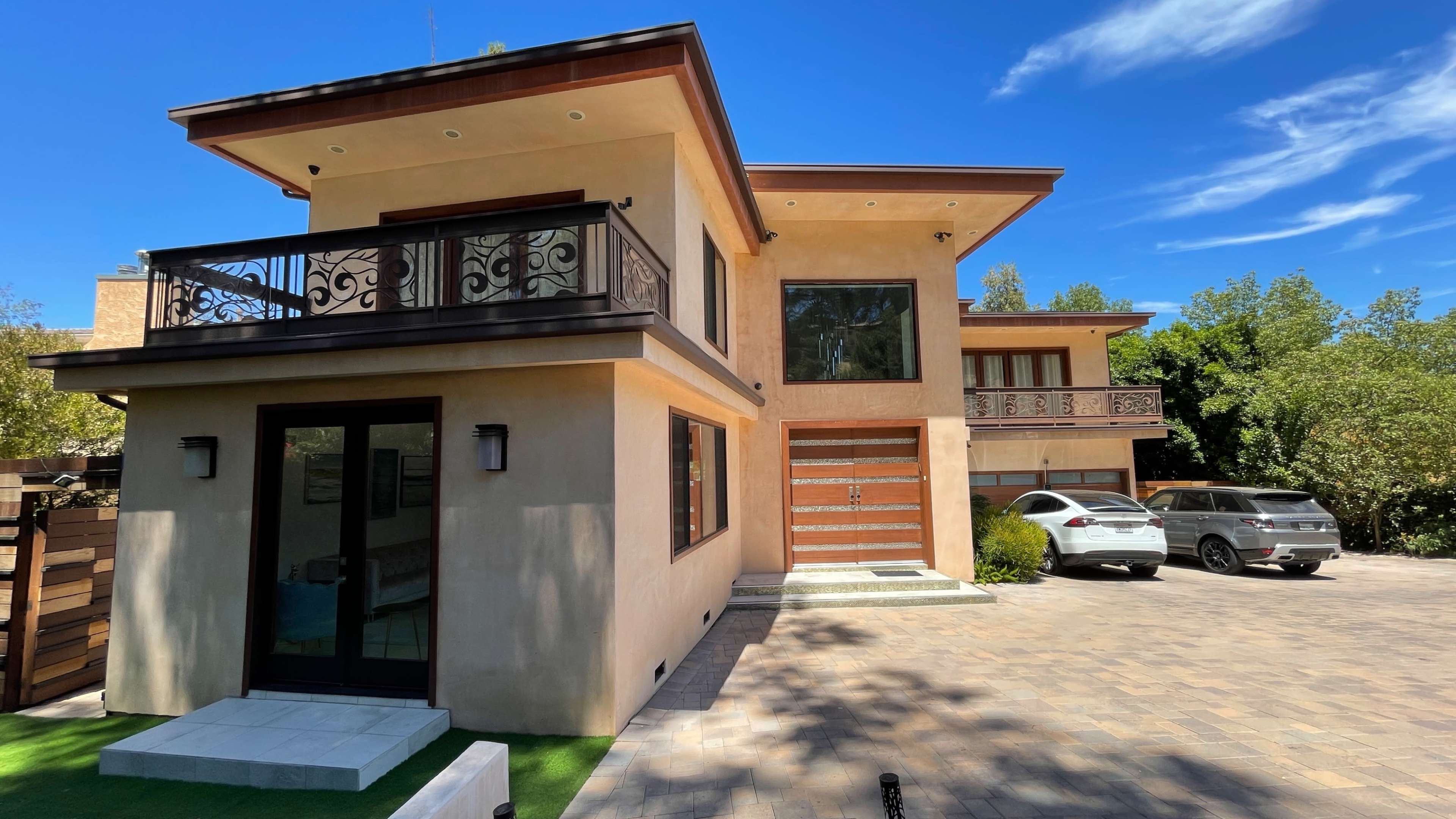 The image shows a modern two-story house with a wooden door, large windows, and a driveway featuring two parked cars.