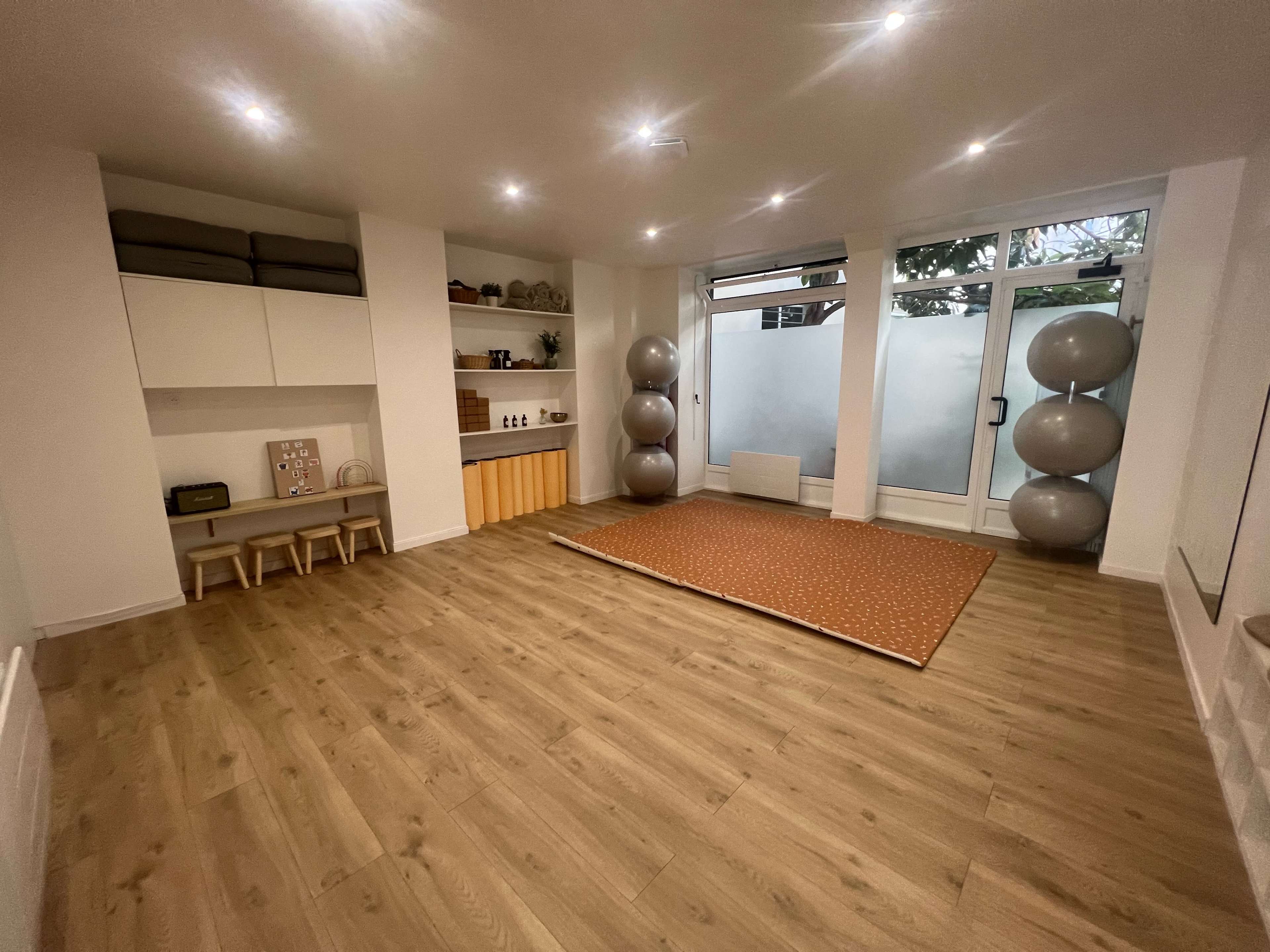 The image shows a spacious room with wooden flooring, equipped with shelves, a seating area, and exercise balls near a large window.