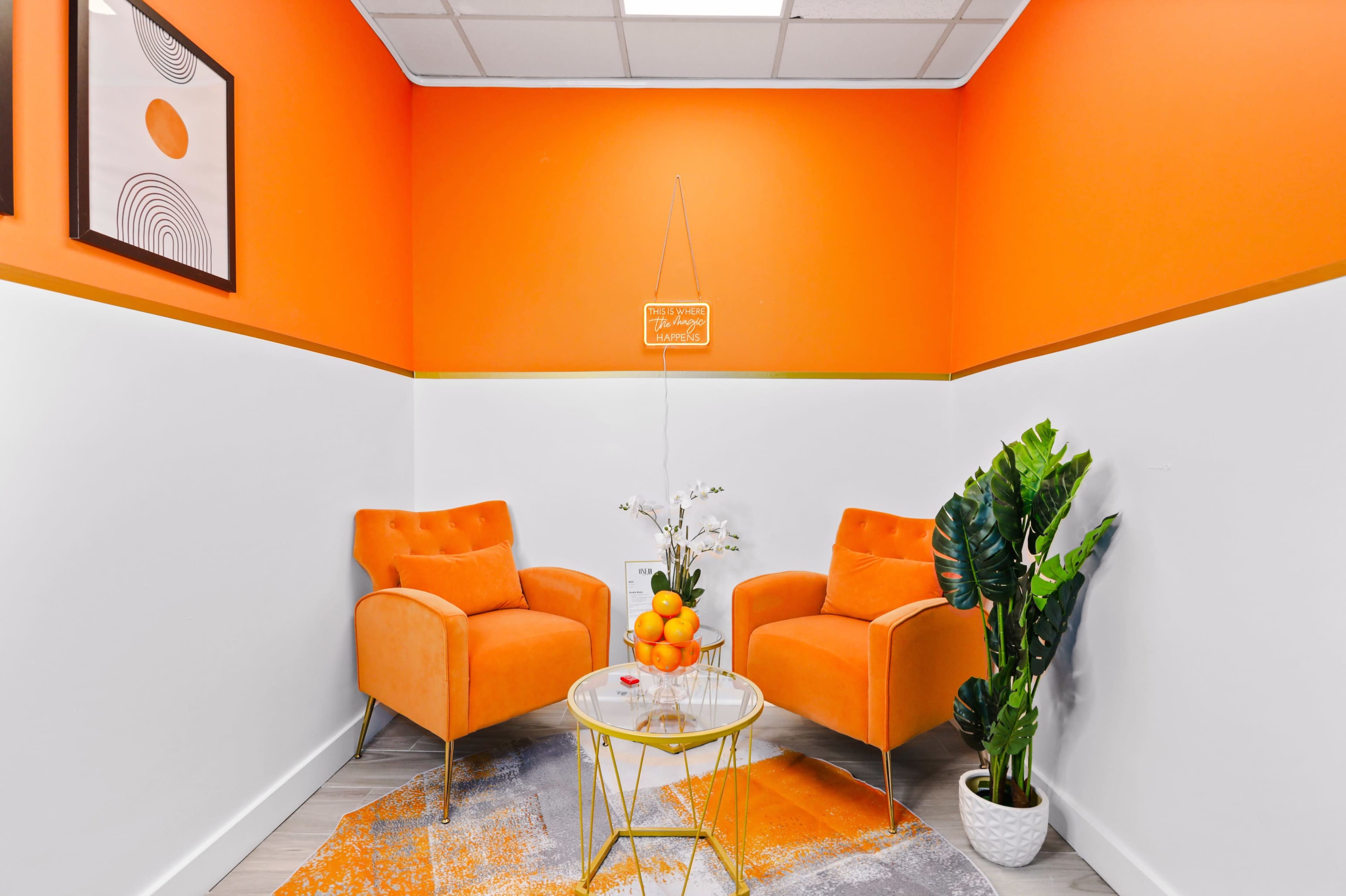 A sunlit corner with two orange chairs, a small glass table with a bowl of oranges, and a potted plant against a bright orange wall.