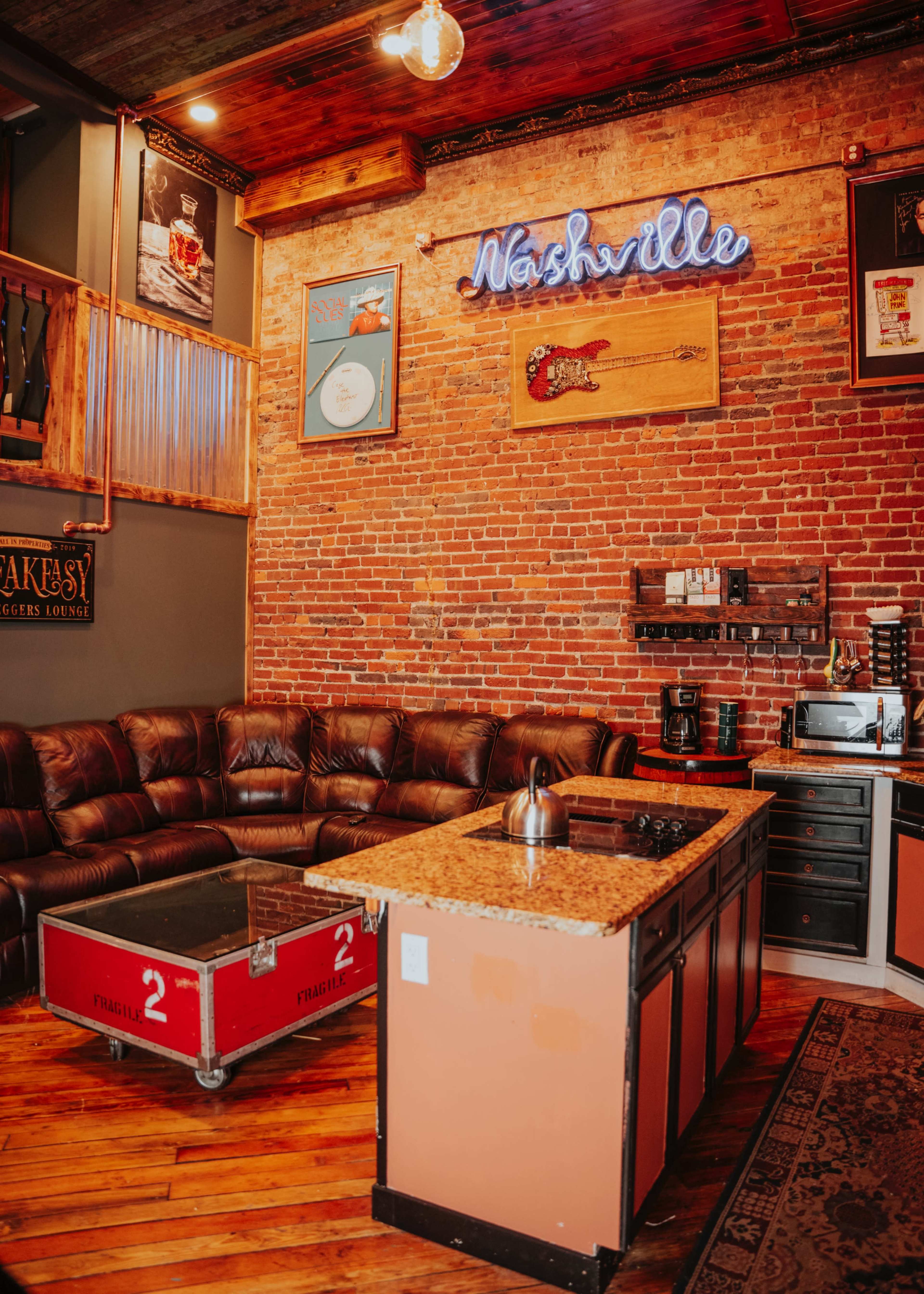 The image shows a cozy lounge area featuring a leather sofa, a kitchen island with a granite countertop, and a brick wall adorned with a neon sign reading "Nashville."