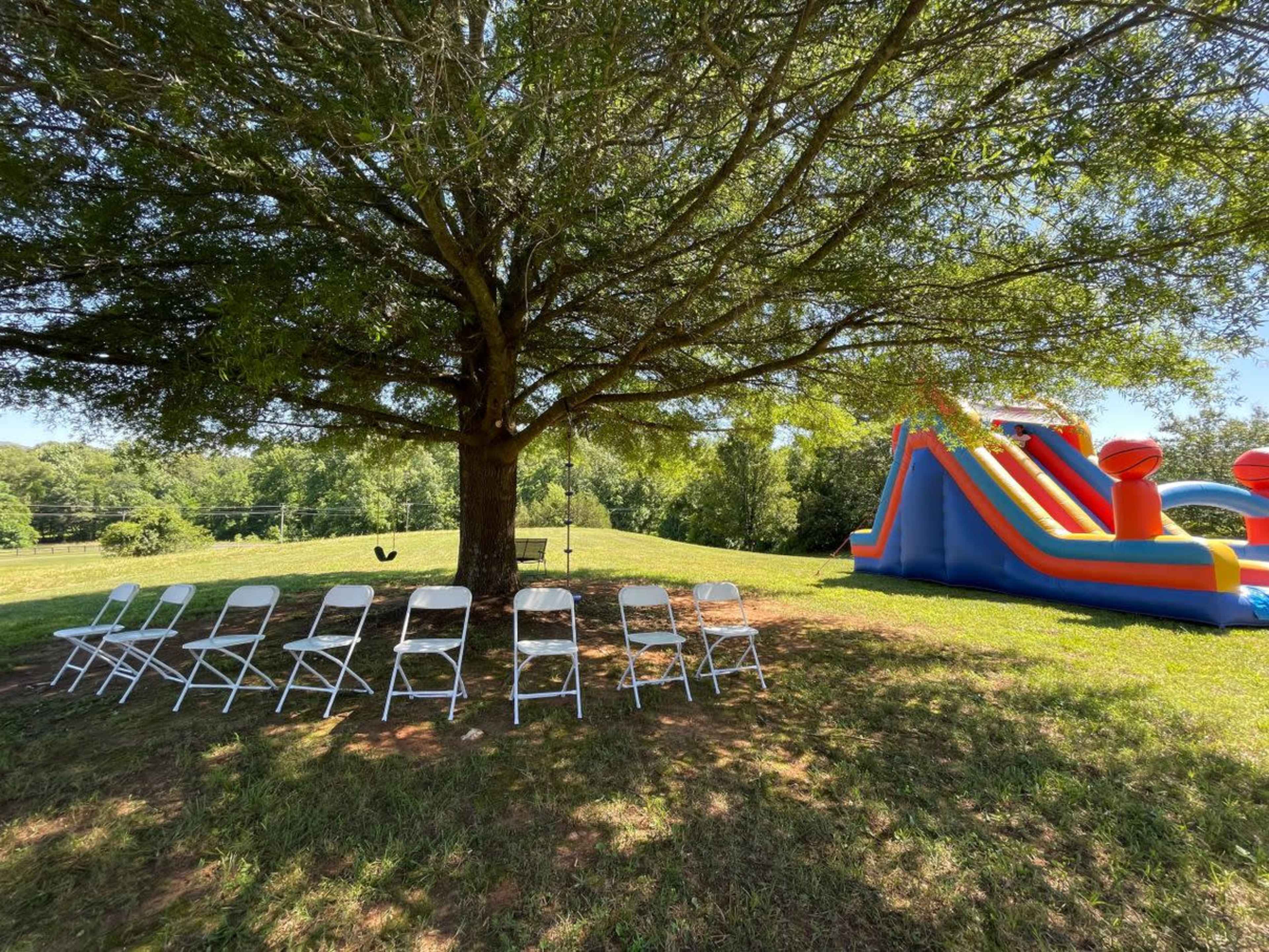 A row of white chairs is arranged under a large tree next to a colorful inflatable slide and bounce house in a grassy area.