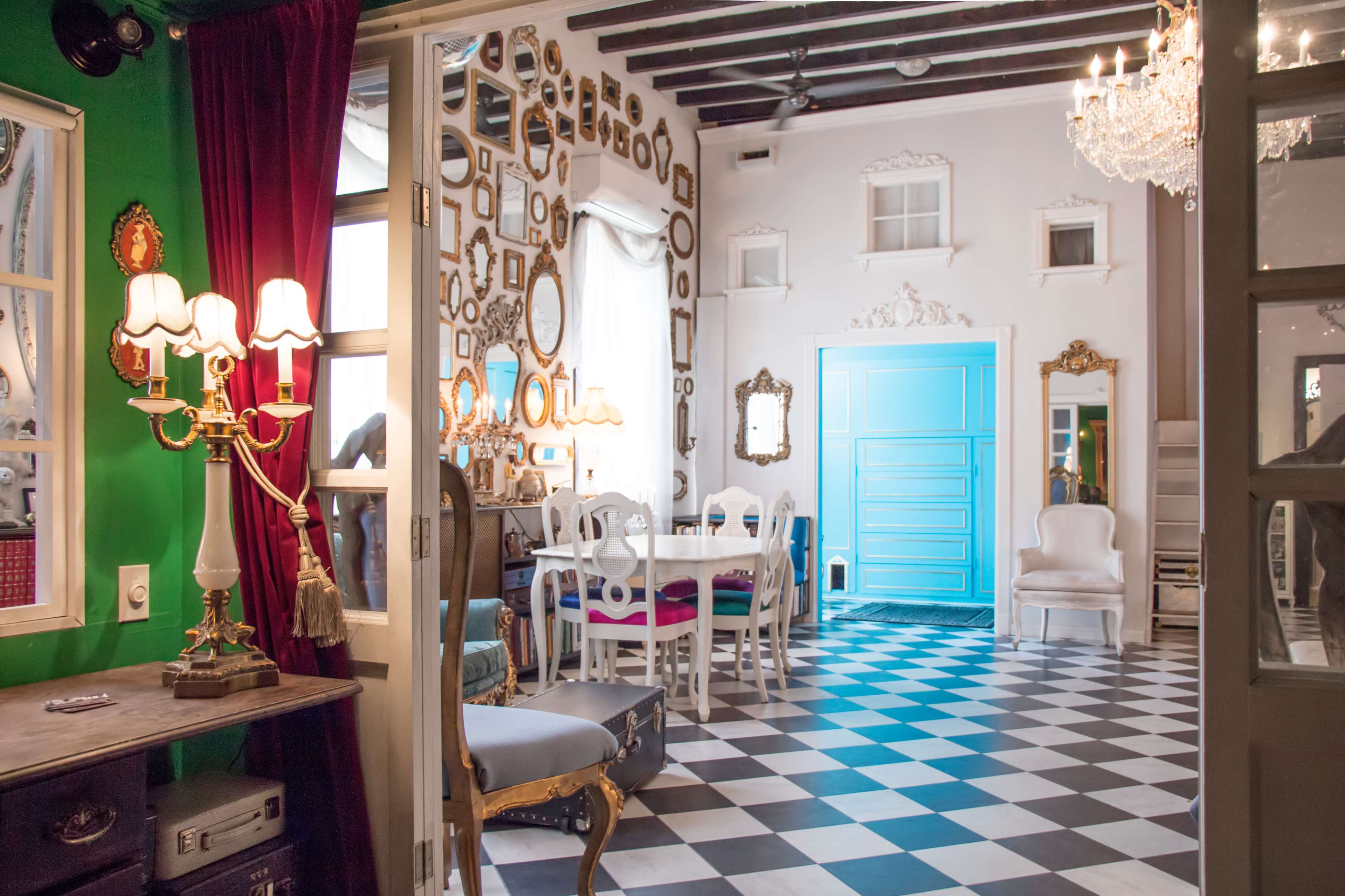 A brightly decorated room with a checkered floor, walls adorned with framed mirrors, and a dining area featuring white furniture and a vibrant blue door.