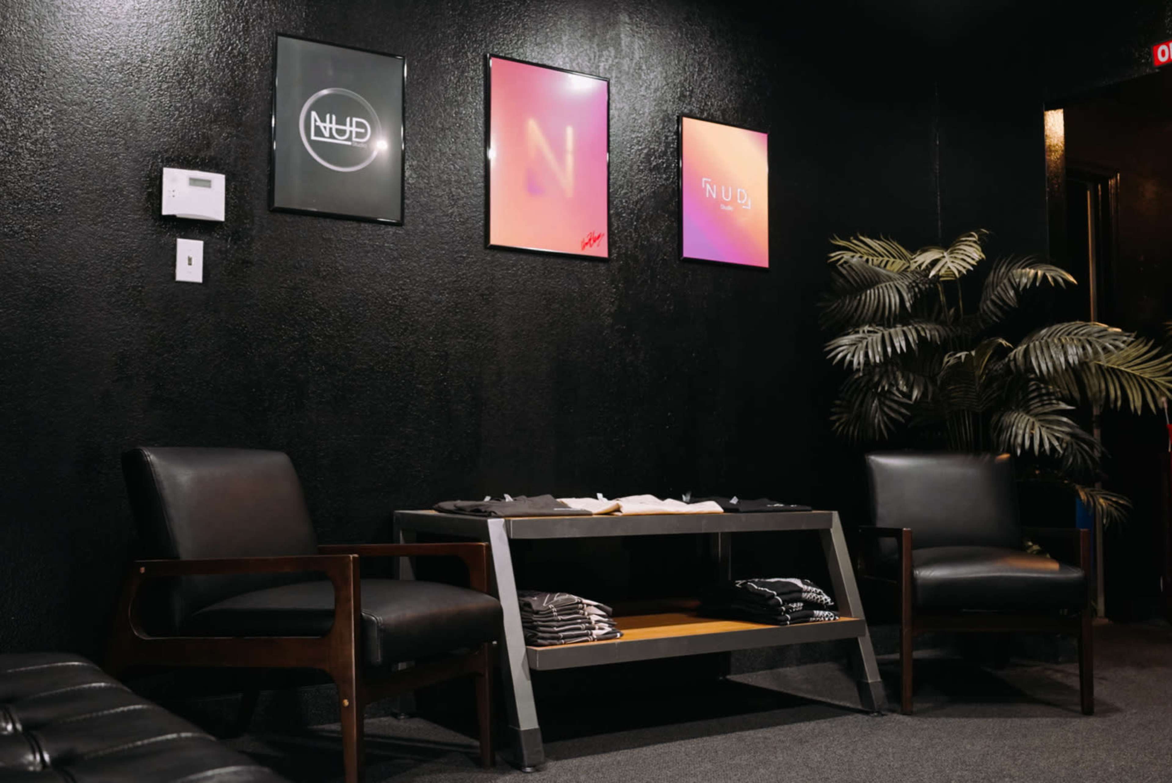 A modern waiting area with two dark chairs, a central table, and three framed artworks on a black wall, alongside a potted plant.