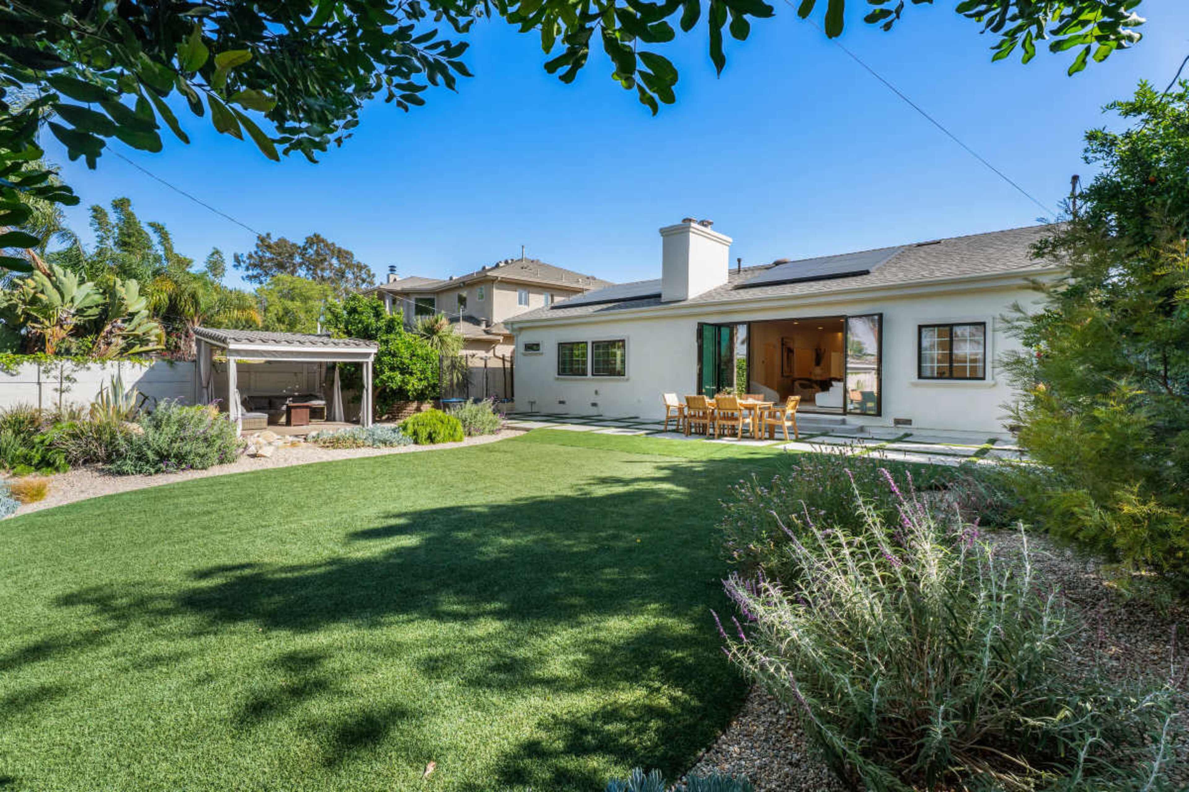 The image depicts a well-maintained backyard featuring a lush green lawn, a seating area on a patio, and a pergola to the side.