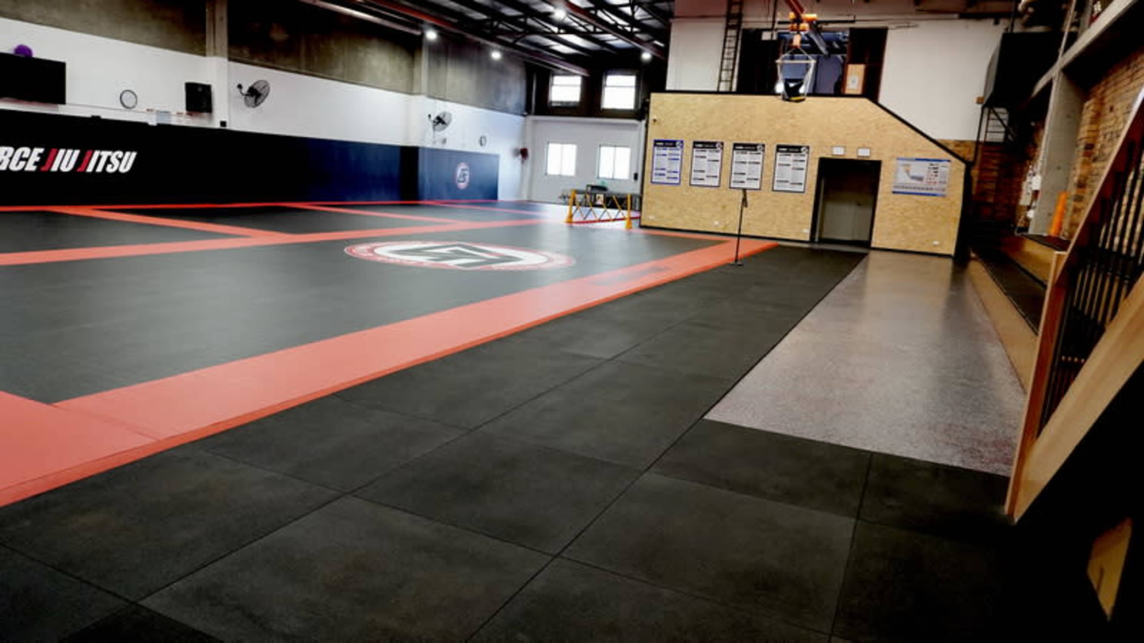 The image shows a spacious martial arts training area with black and red mats, equipped with posters and a staircase in the background.