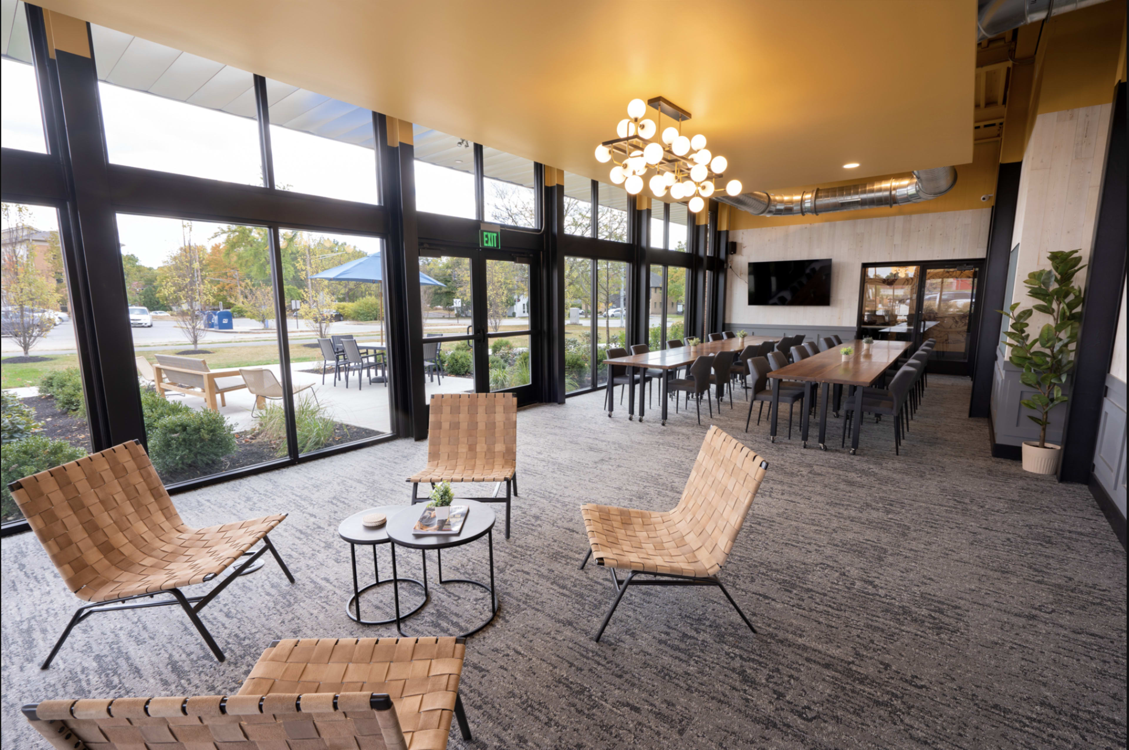 The image shows a modern interior space with large windows, featuring four woven chairs around a small table and a long communal dining table in the background.
