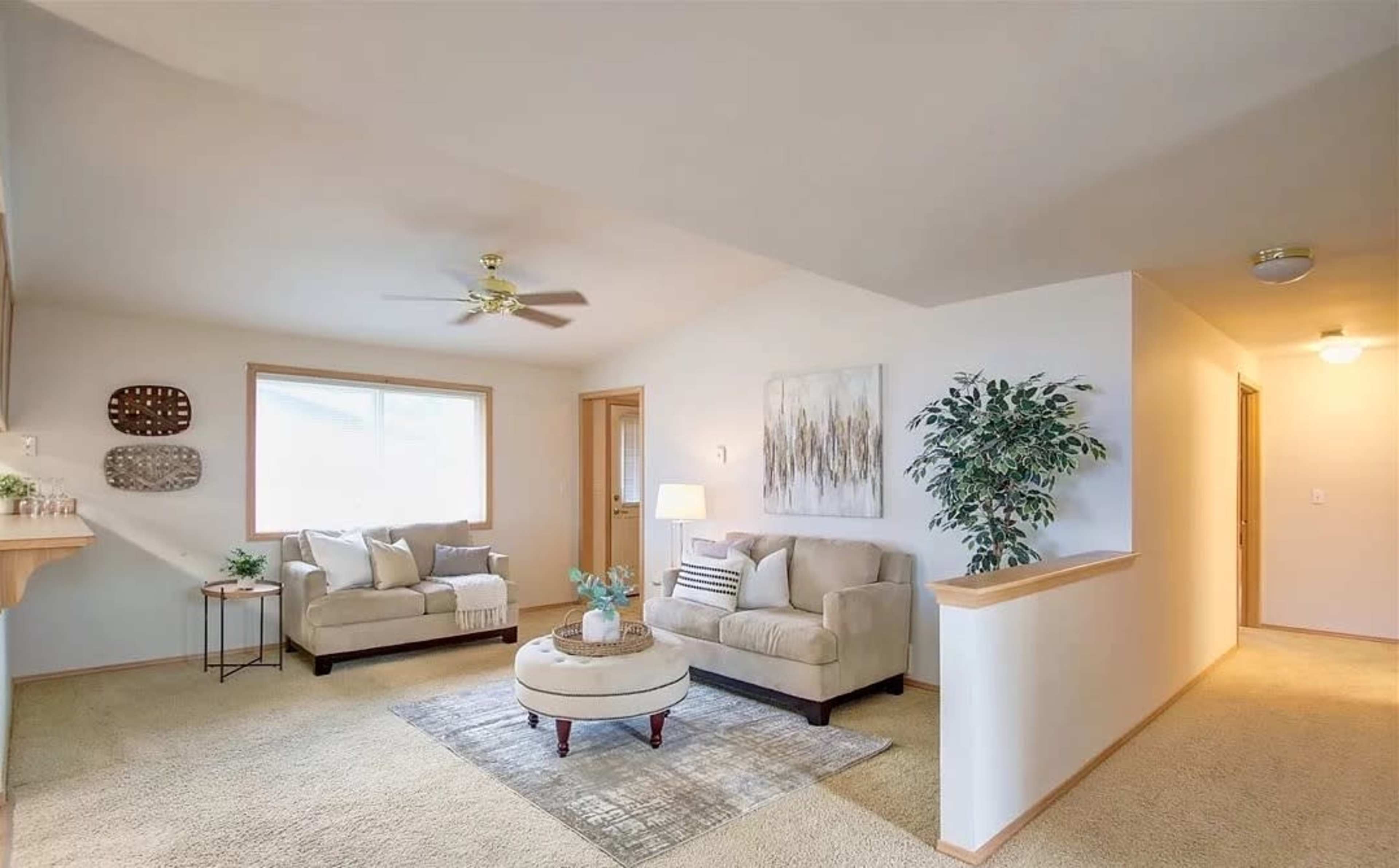 A well-lit living room features two beige sofas, a round coffee table, and a potted plant, with a door leading to a hallway.