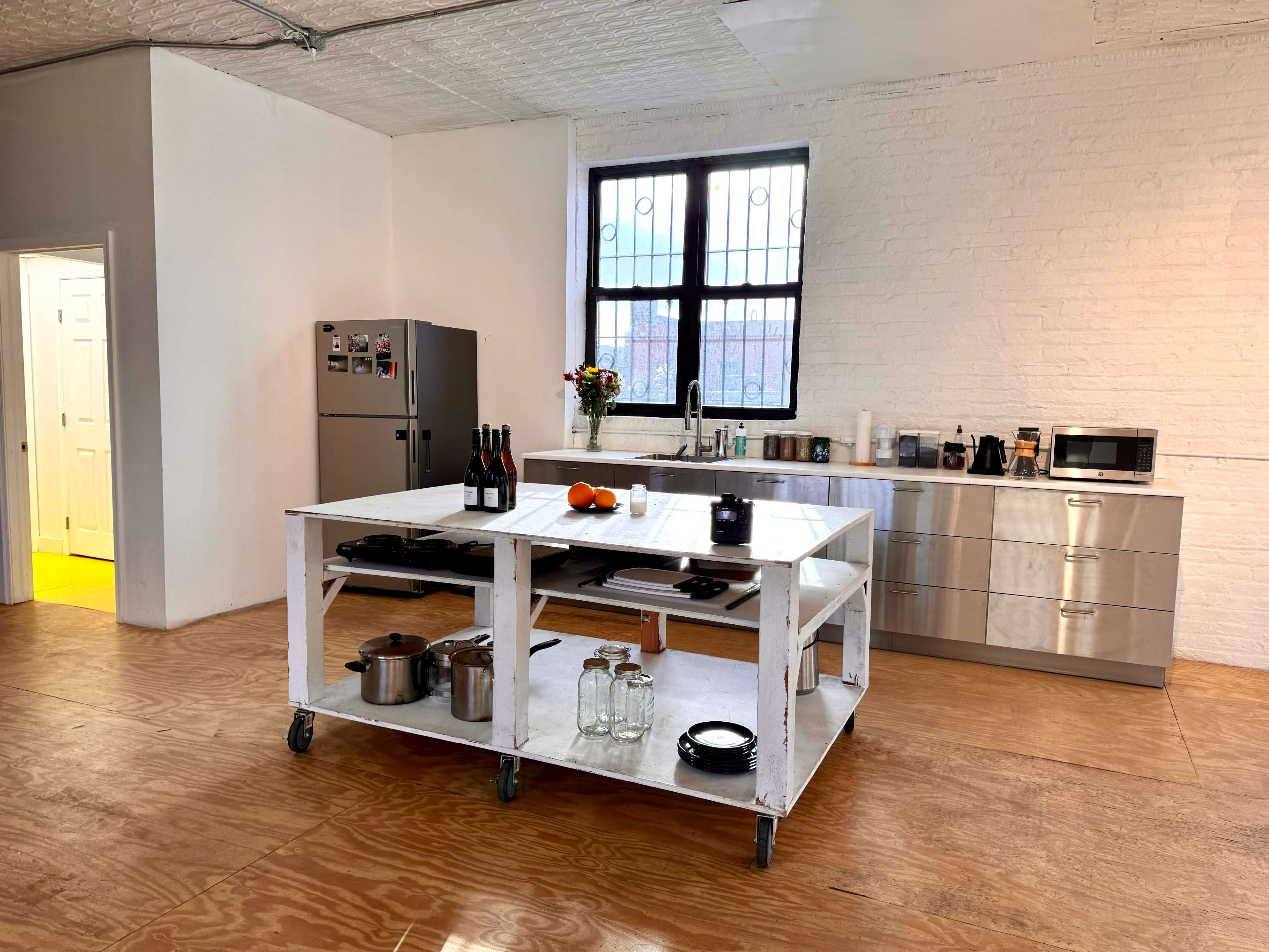 The image shows a spacious kitchen with a central island on wheels, stainless steel cabinetry, a refrigerator, and various kitchen appliances.