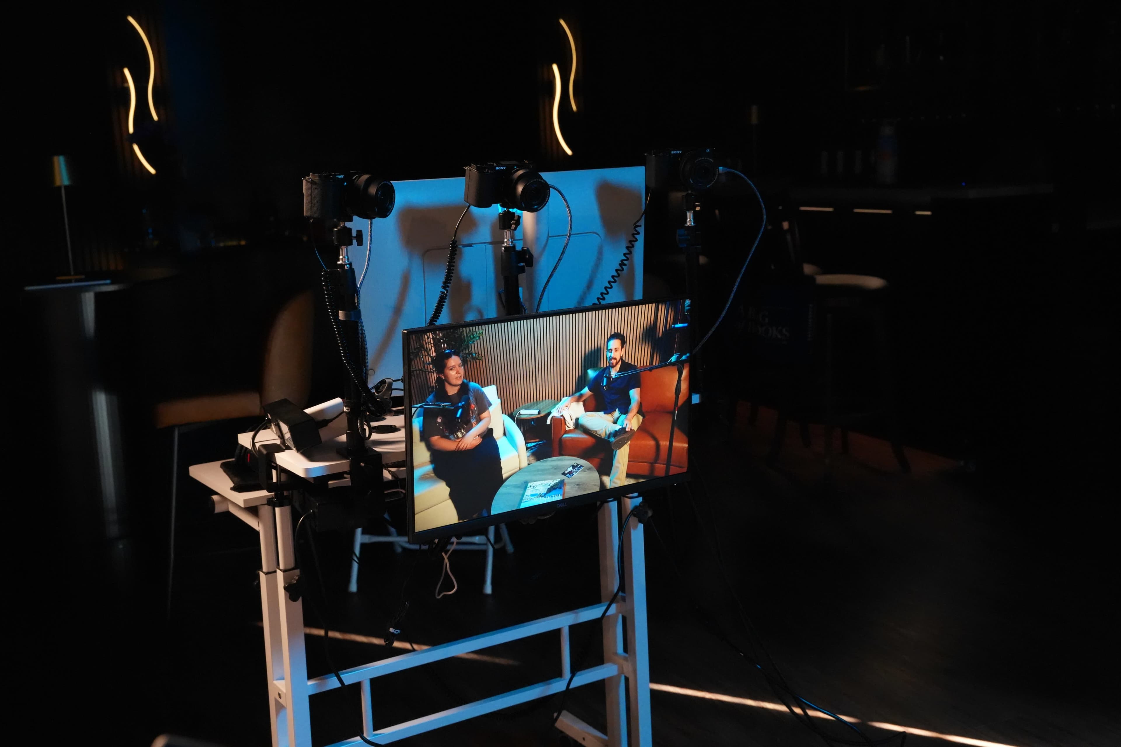 A monitor displays two people sitting on a couch, surrounded by cameras and equipment in a dimly lit room.