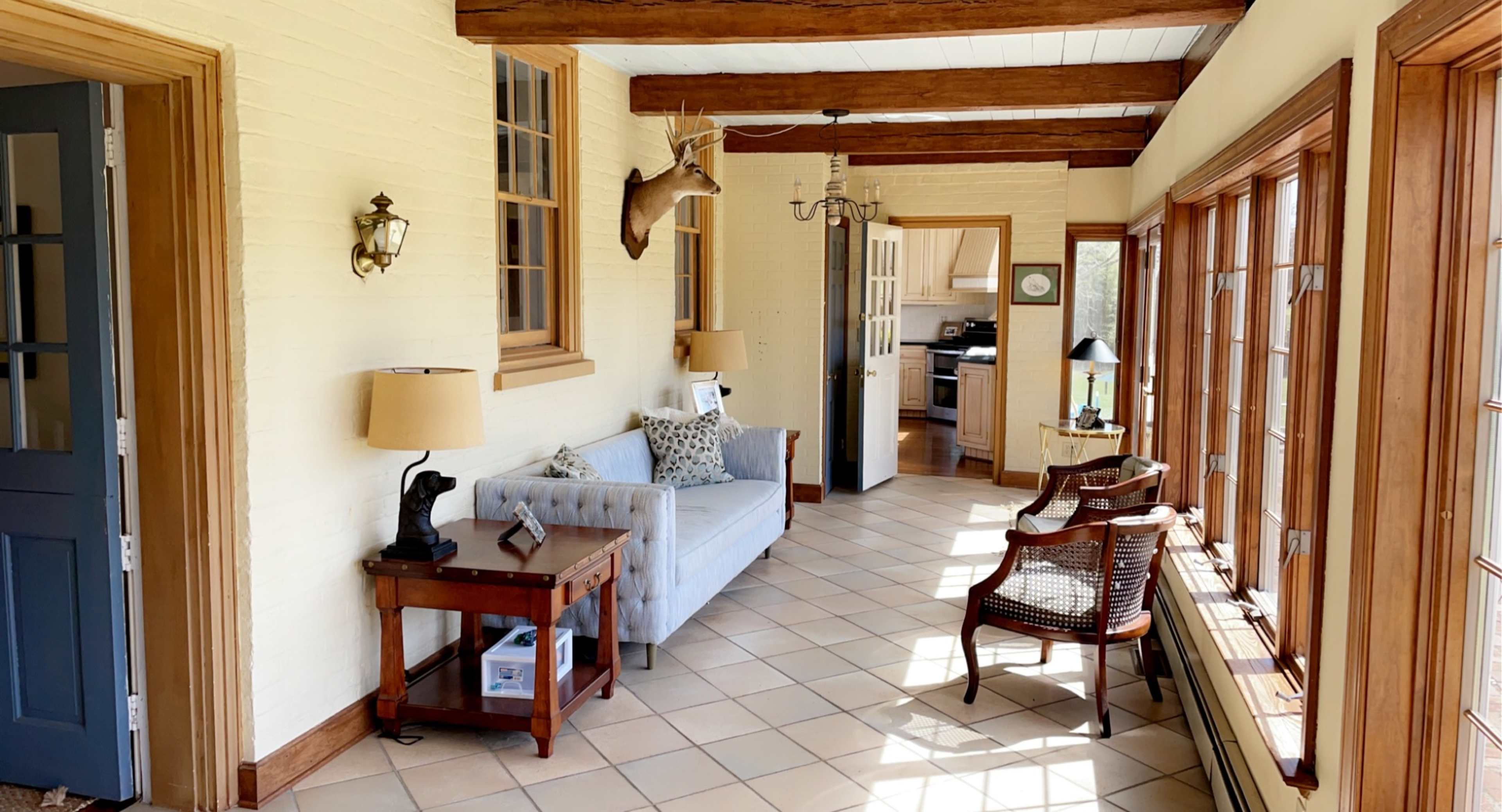 The image shows a sunlit hallway with a blue sofa, wooden beams overhead, and large windows, leading to a kitchen.