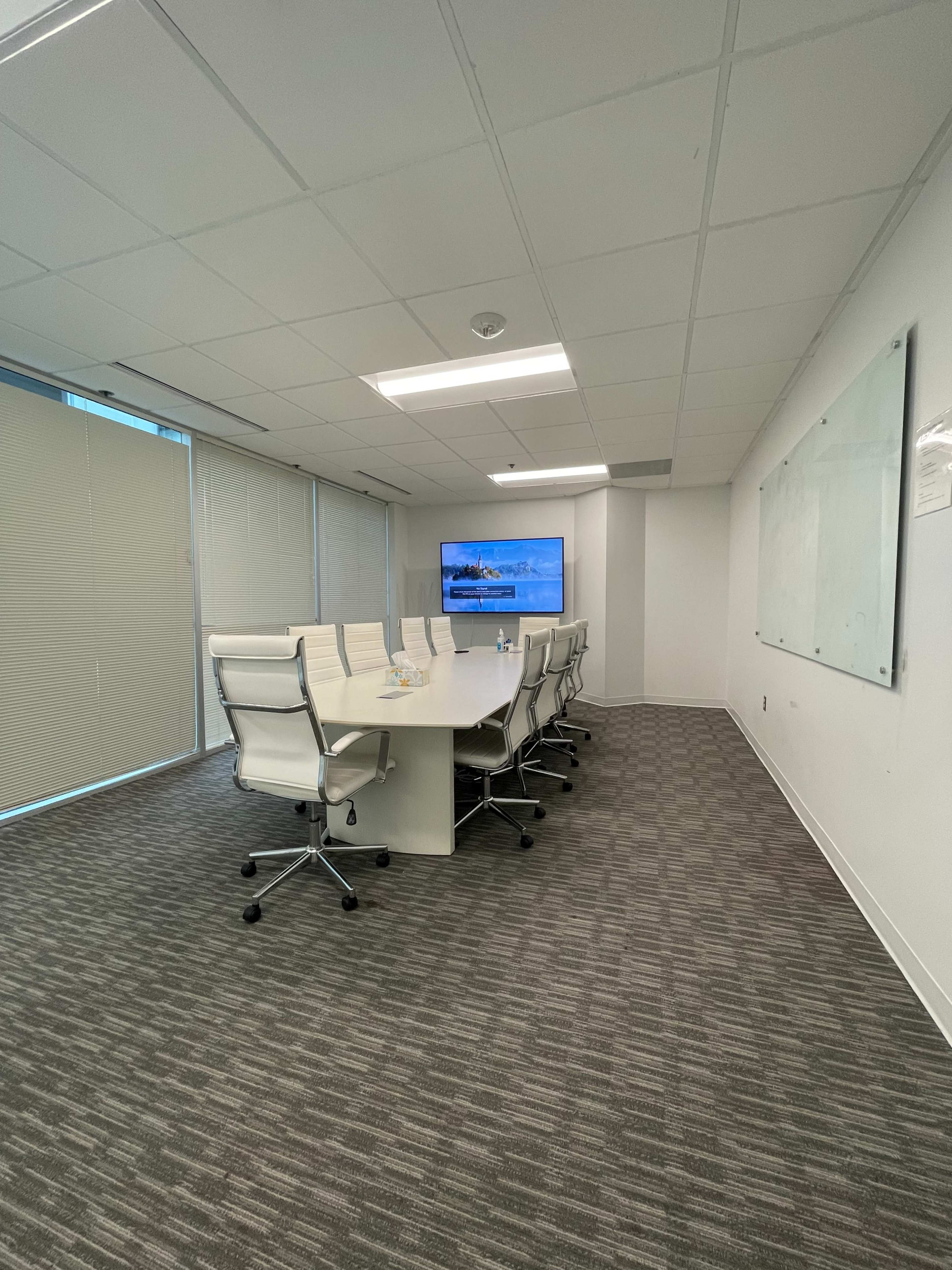 A conference room features a long white table with eight chairs, a large screen on the wall, and blinds covering the windows.