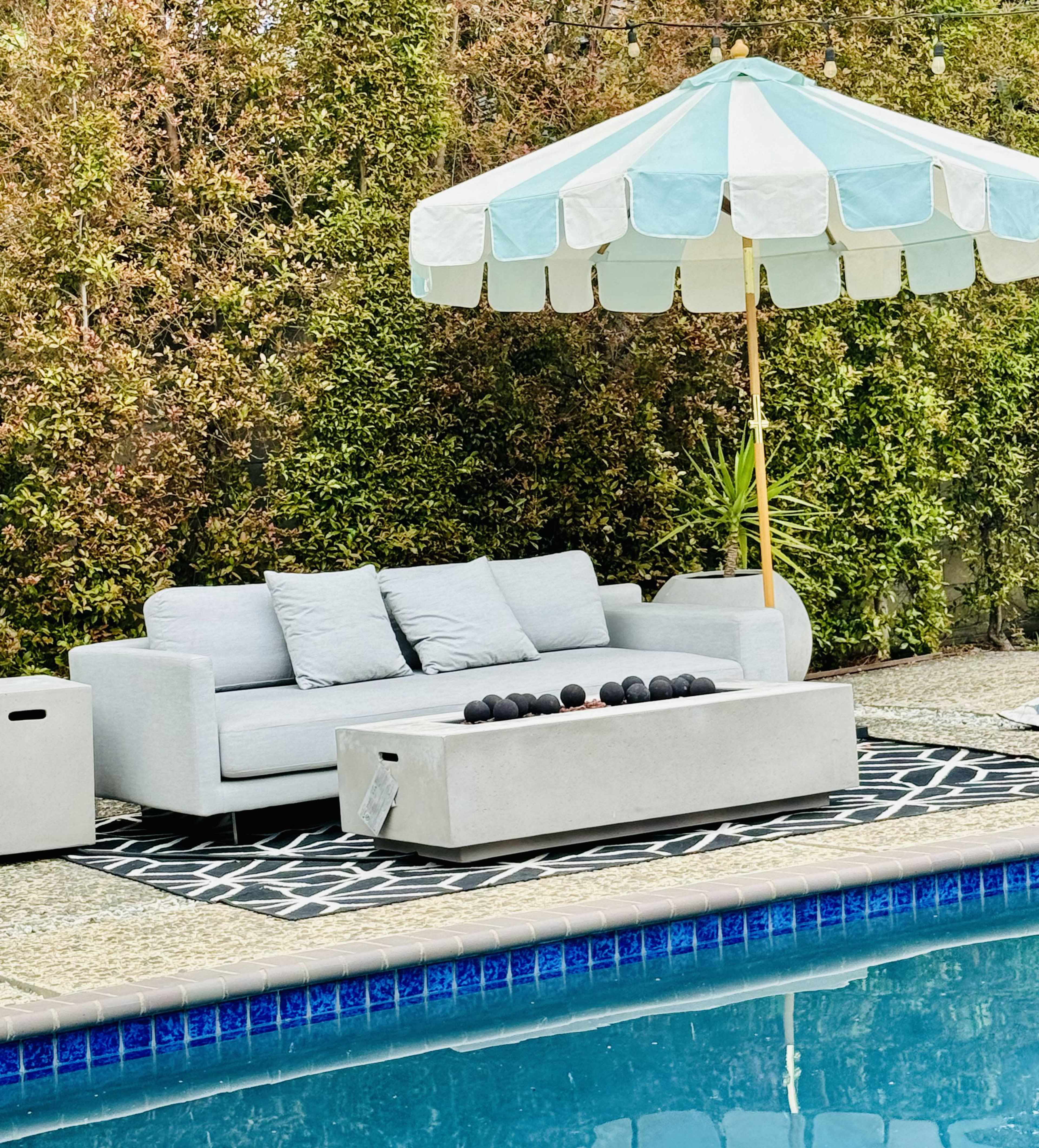 A modern outdoor seating area with a gray sofa, a concrete coffee table, and a large striped umbrella beside a swimming pool.