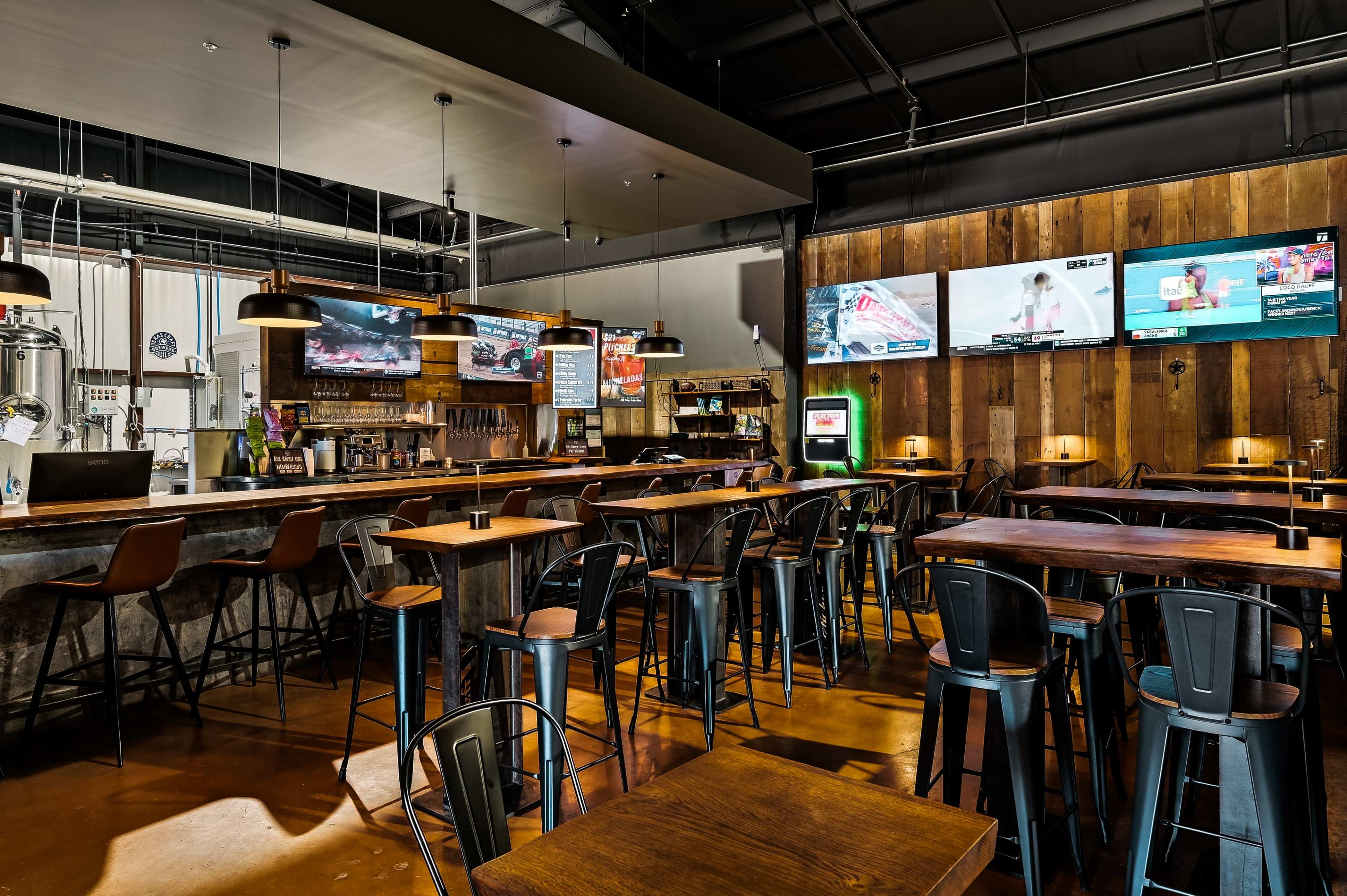 The image shows a modern bar area with a wooden accent wall, high-top tables, and multiple televisions displaying various programs.