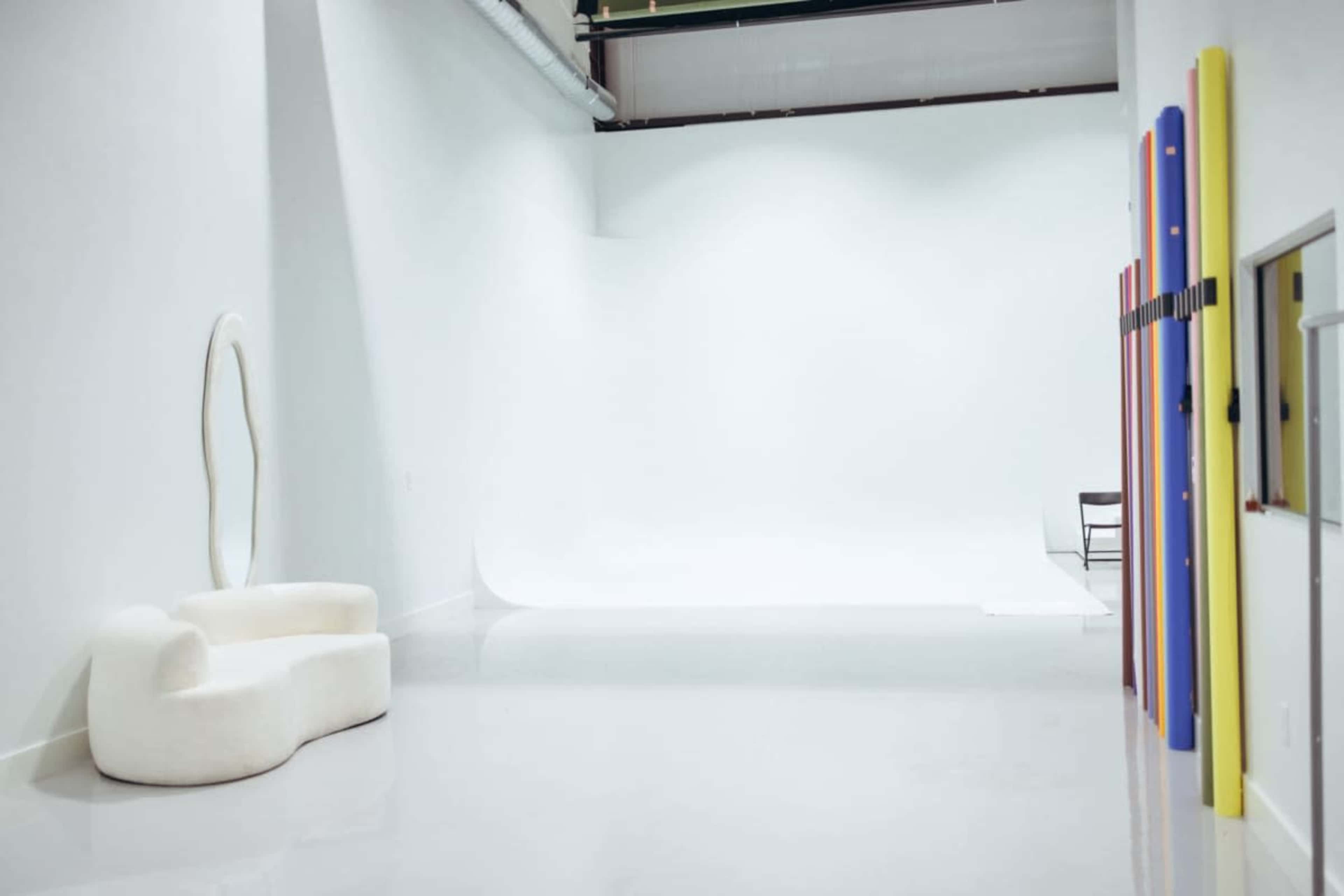 The image depicts a bright, minimalist studio space featuring a curved white couch, a blank backdrop, and colorful vertical panels along one wall.