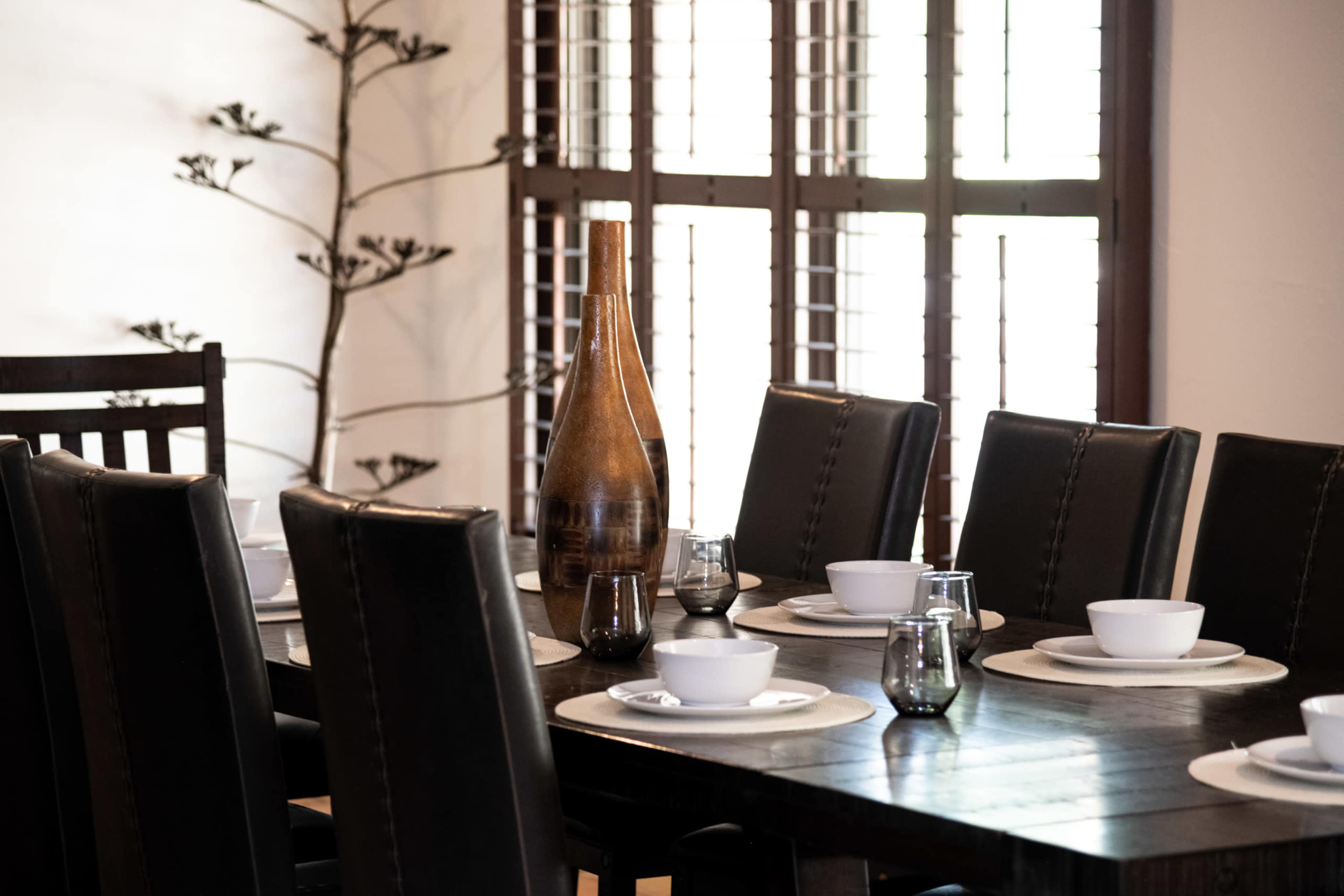 A long dining table is set with white bowls and glasses, with a decorative brown bottle at the center and a minimalist plant in the background.