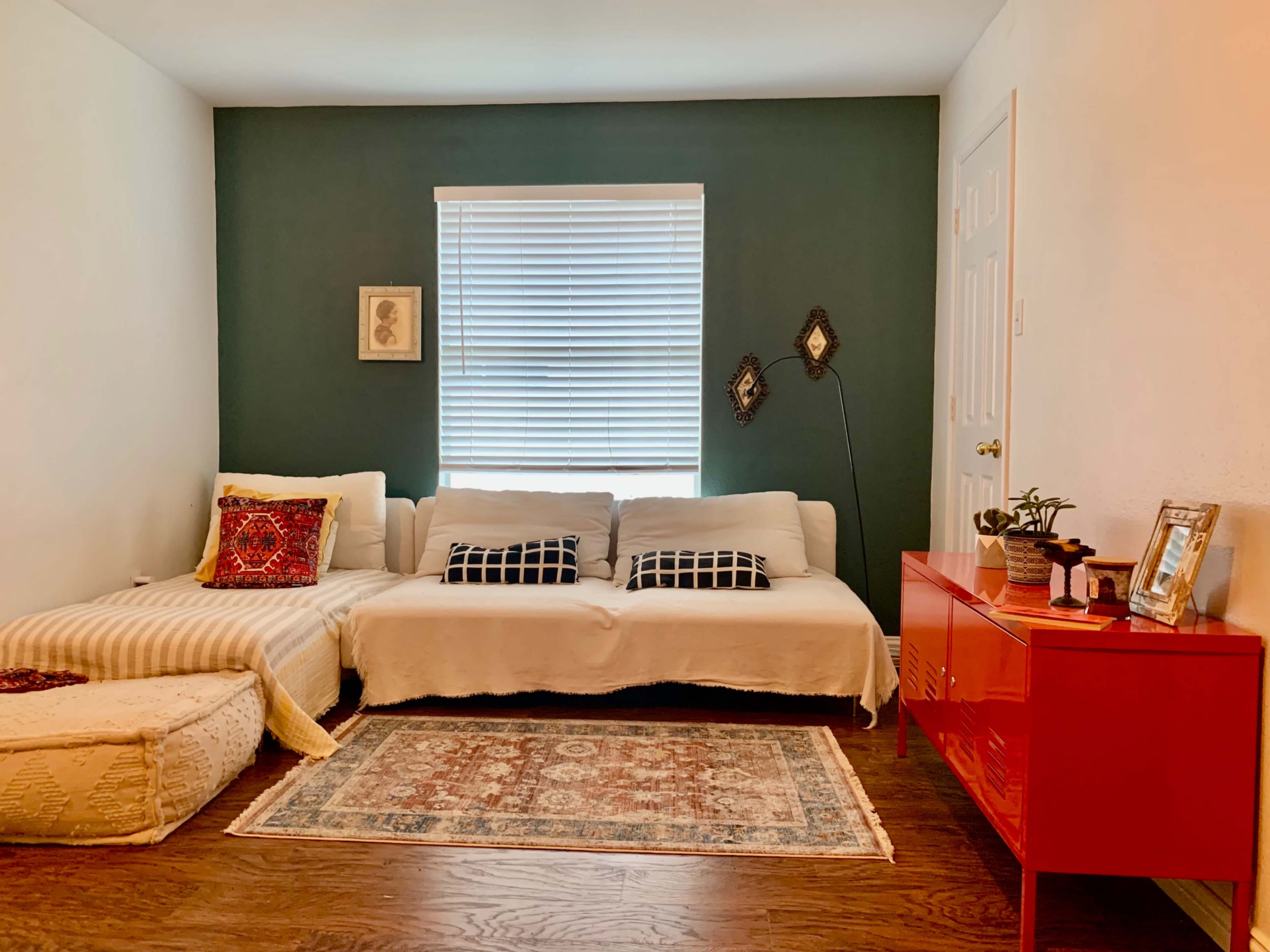 A living room with a white sofa adorned with decorative pillows, a red cabinet, a patterned rug, and a window with blinds.