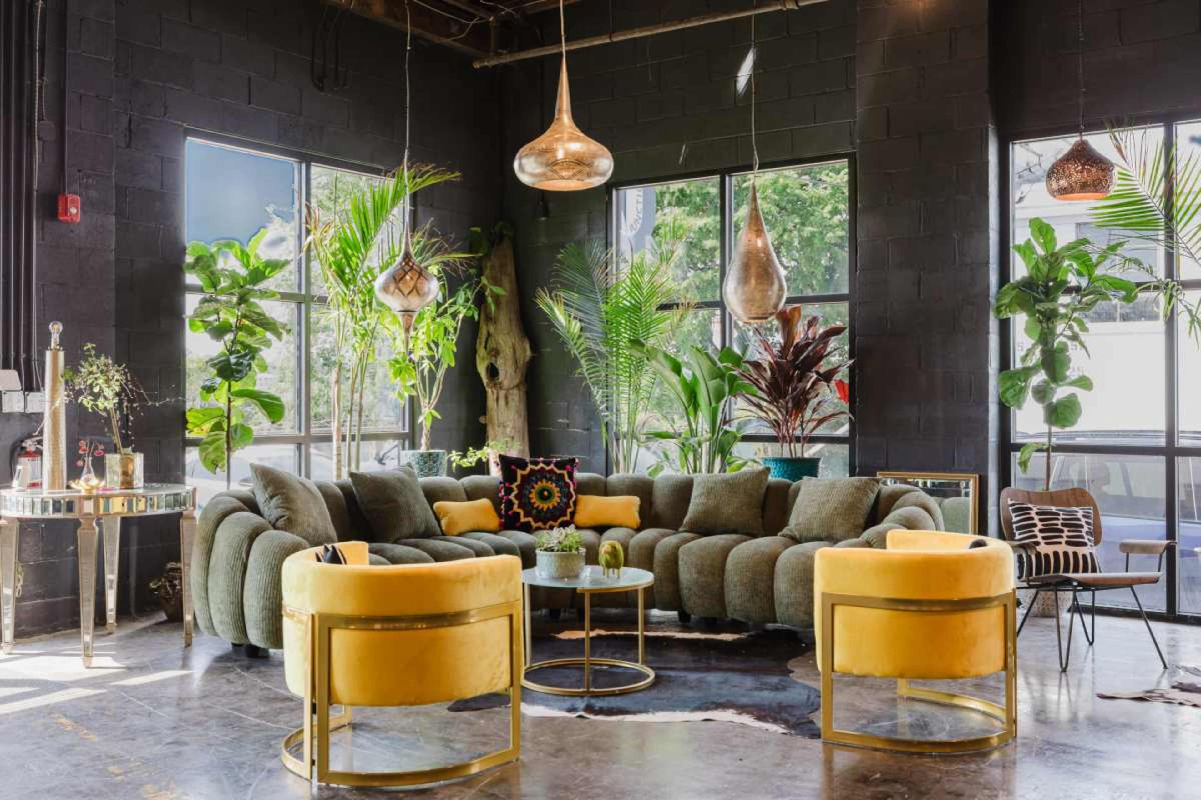 The scene features a modern lounge area with a curved green sofa, two yellow accent chairs, and large windows surrounded by various indoor plants.