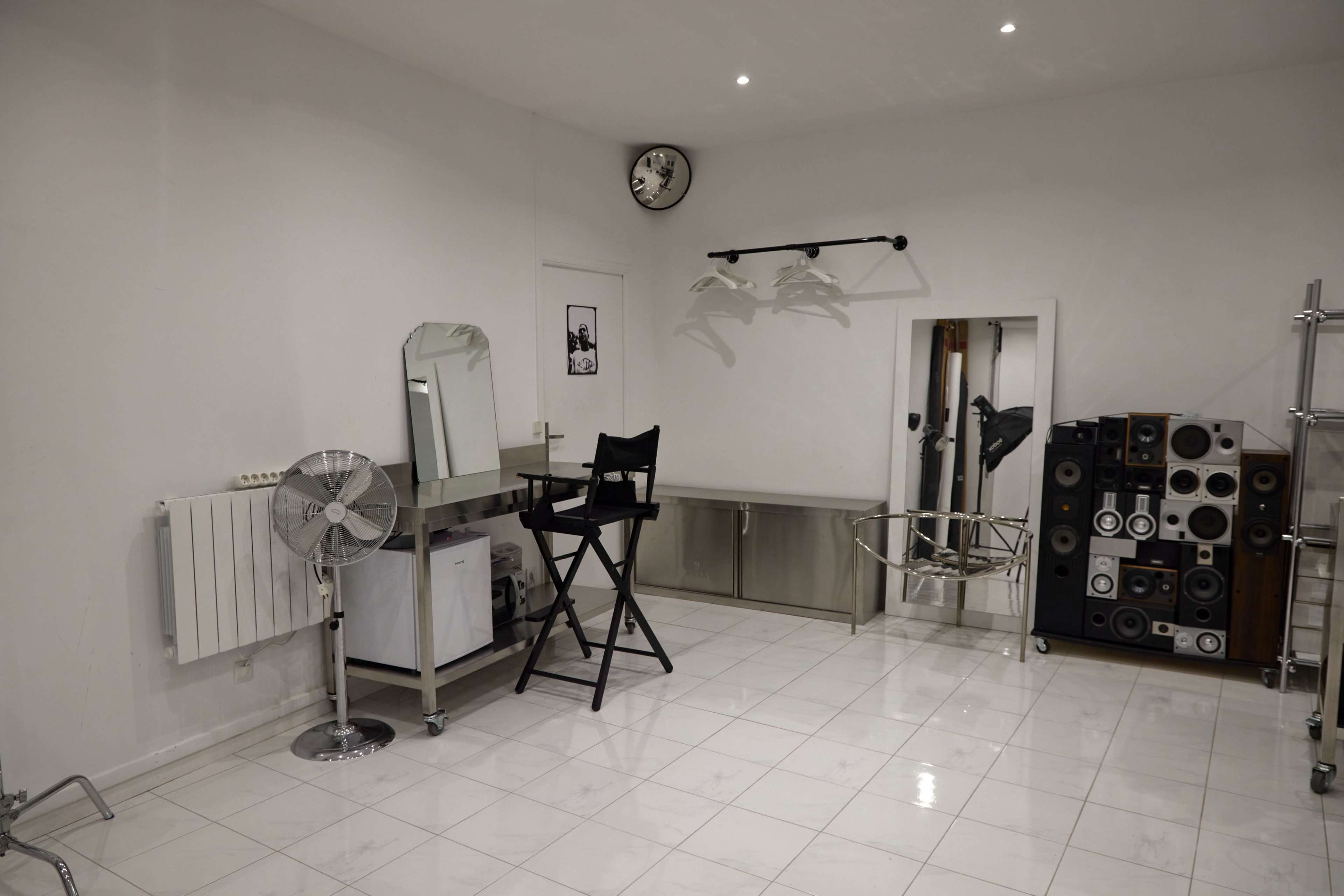 The image shows a minimalist, well-lit room with a chair, a mirror, a fan, and various storage units along the walls.