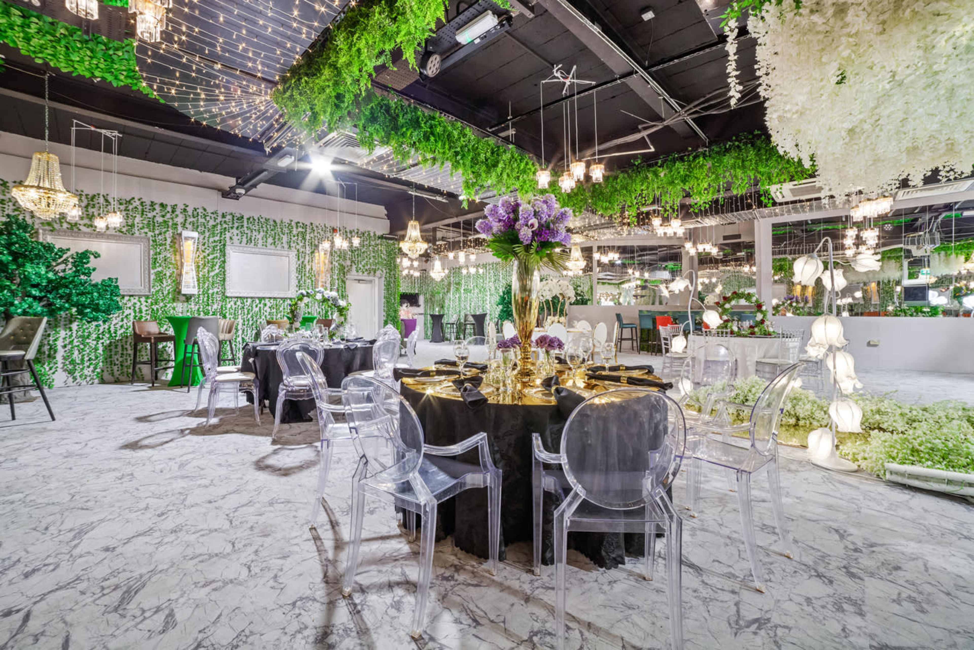 The scene features an elegantly decorated dining space with a black table set for a banquet, surrounded by transparent chairs and adorned with greenery and floral arrangements overhead.