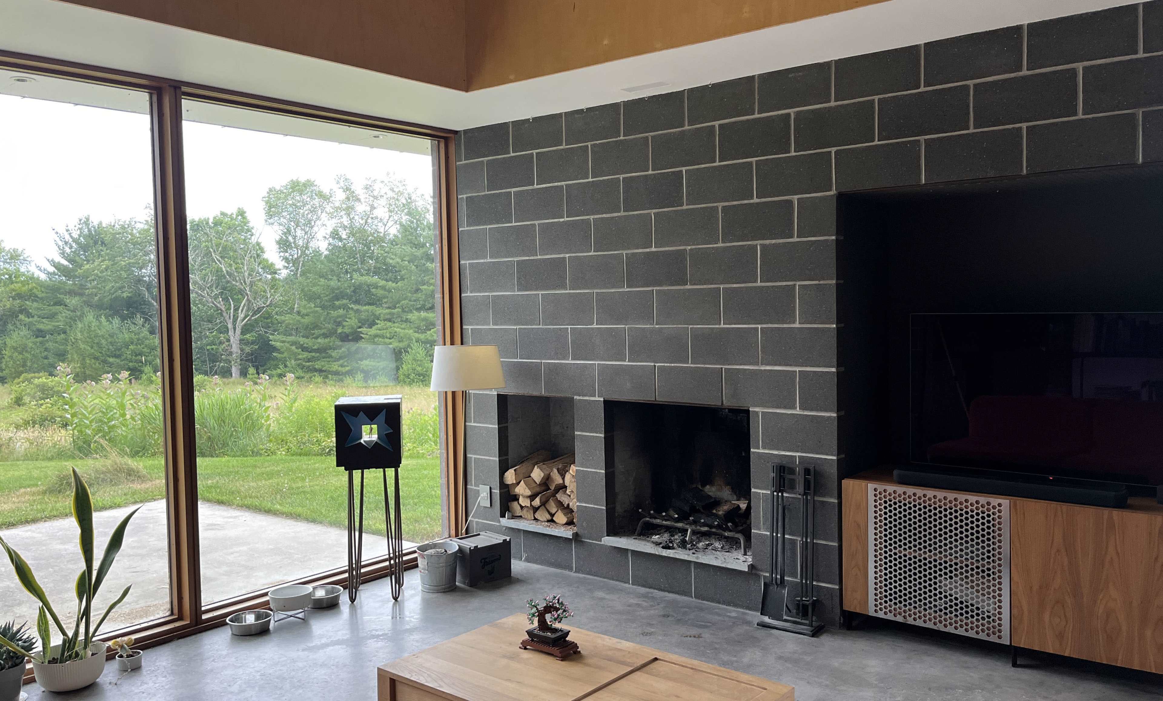 A modern living room features a stone wall with a fireplace, large windows overlooking a grassy area, and minimal furnishings.