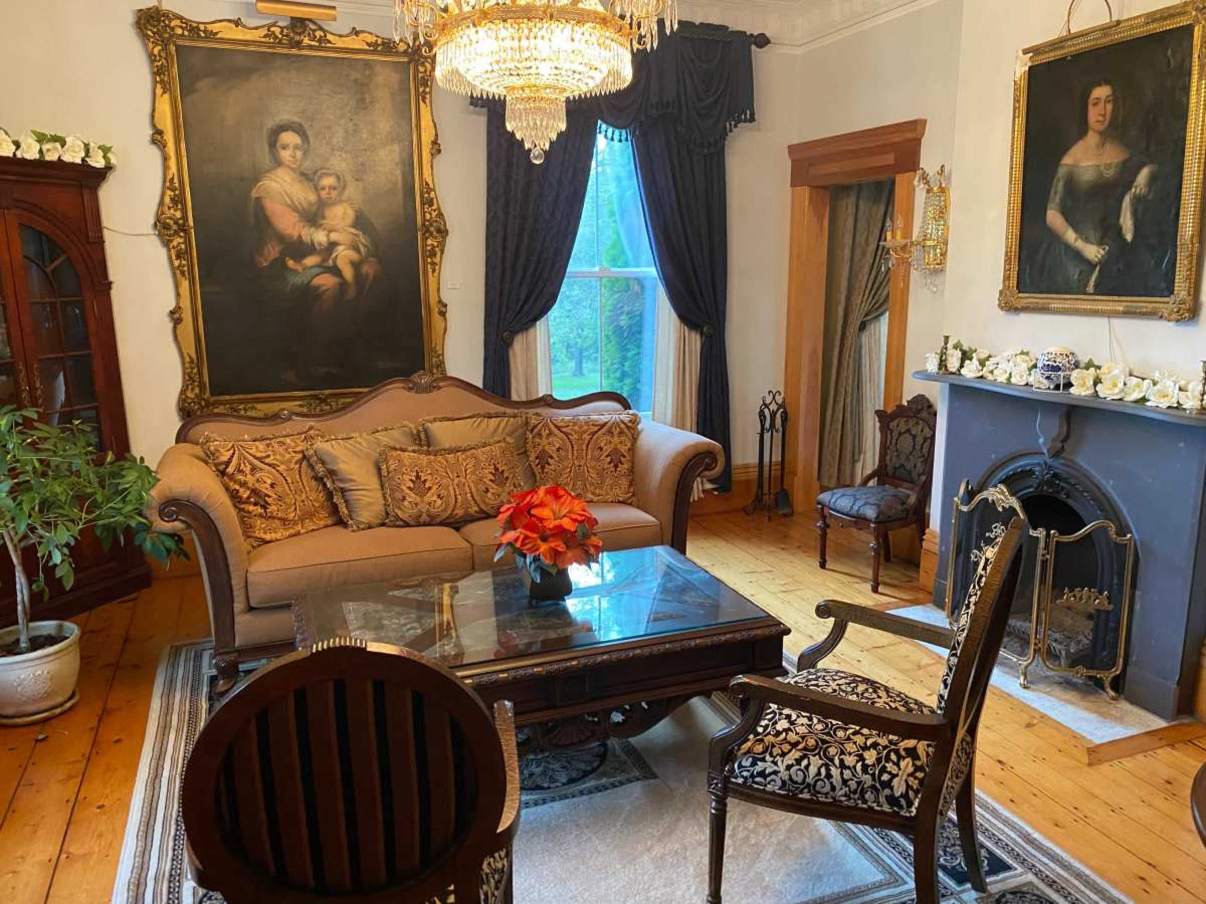 The room features a chandelier, ornate furniture, and framed portraits, including one of a woman, with a large painting above a couch and a fireplace.