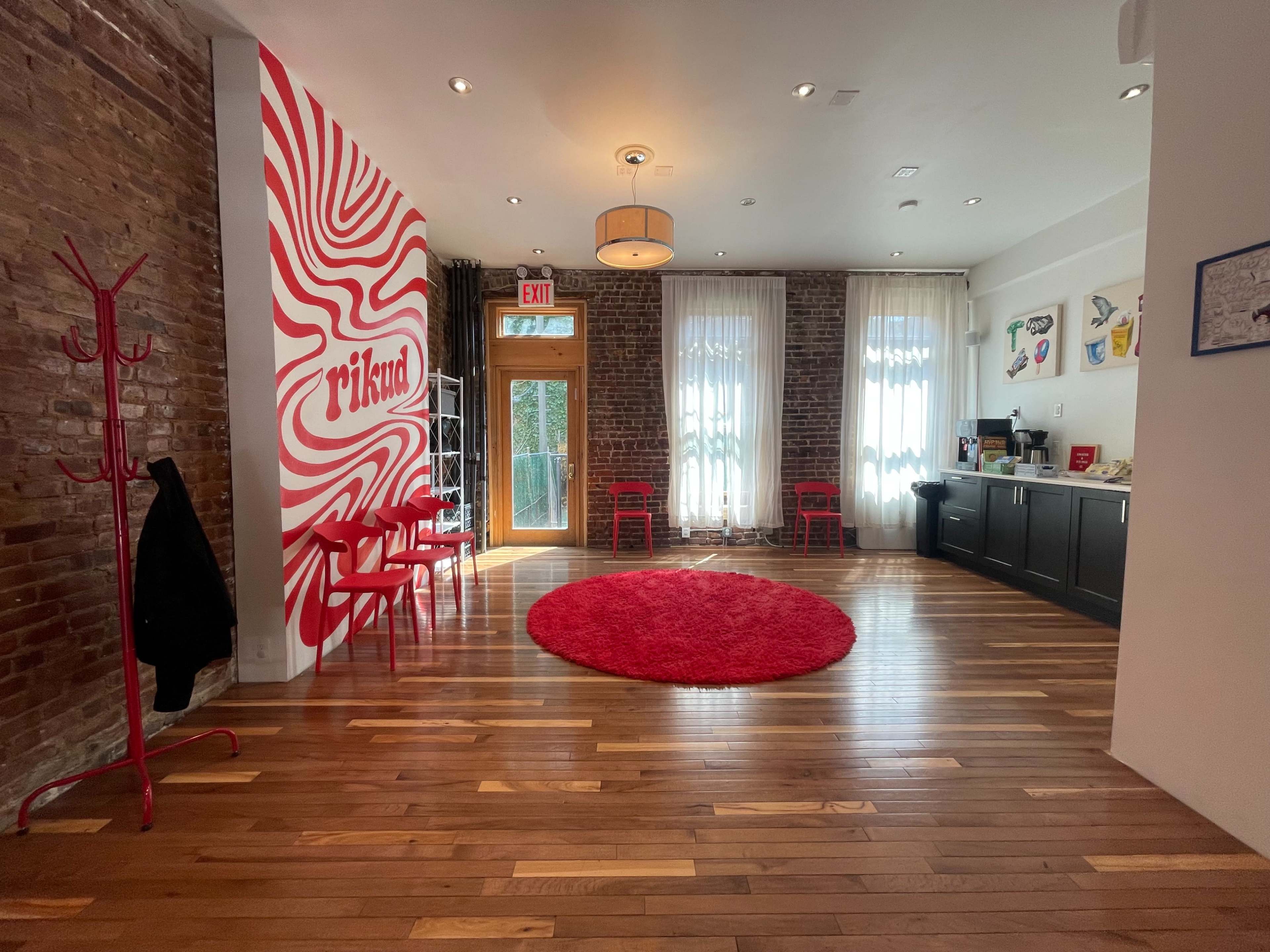 The image shows a bright room with exposed brick walls, a circular red rug on a wooden floor, red chairs, and a white wall featuring a wavy red design.