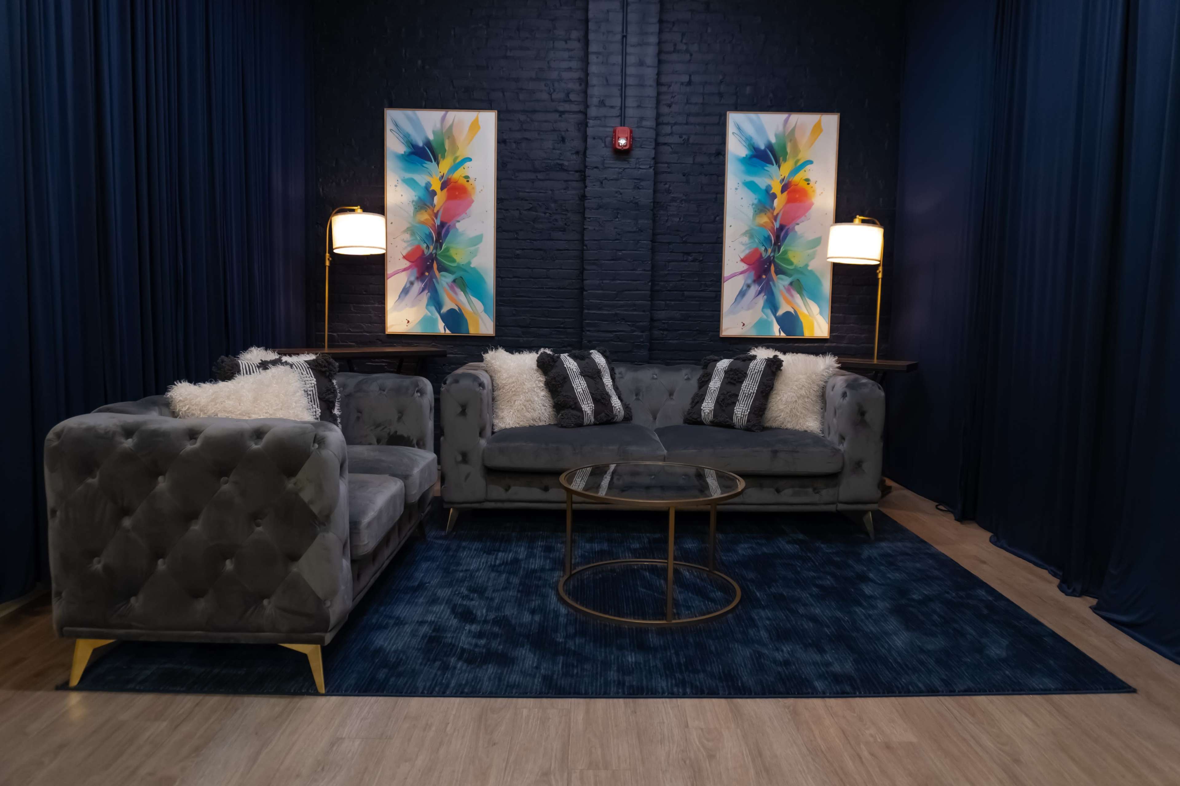 The image shows a cozy lounge area featuring a gray tufted sofa set with decorative pillows, a round coffee table, and vibrant abstract artwork on the walls.