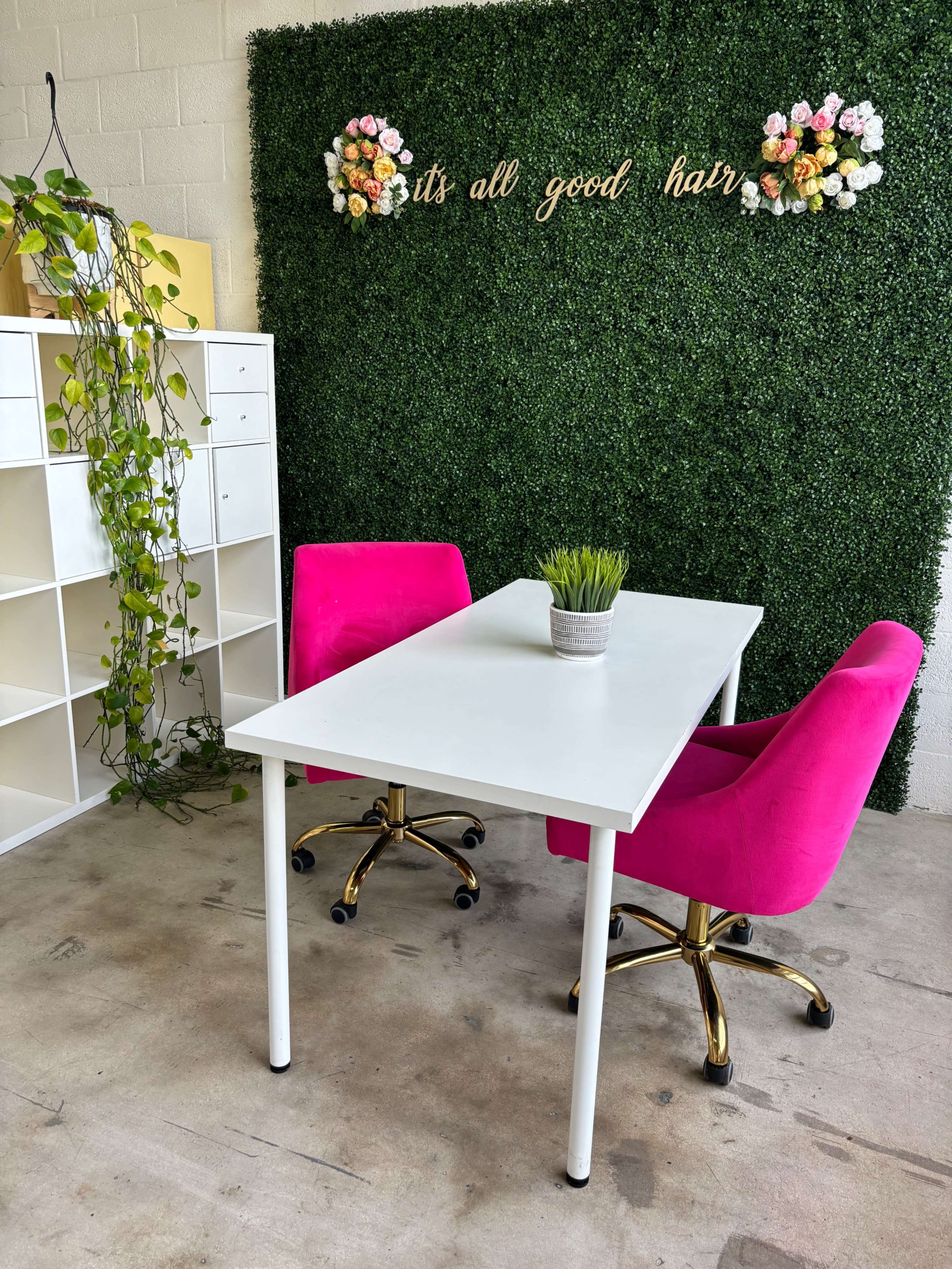 A white table with two pink chairs sits in front of a green wall adorned with flowers and the text "it's all good hair."