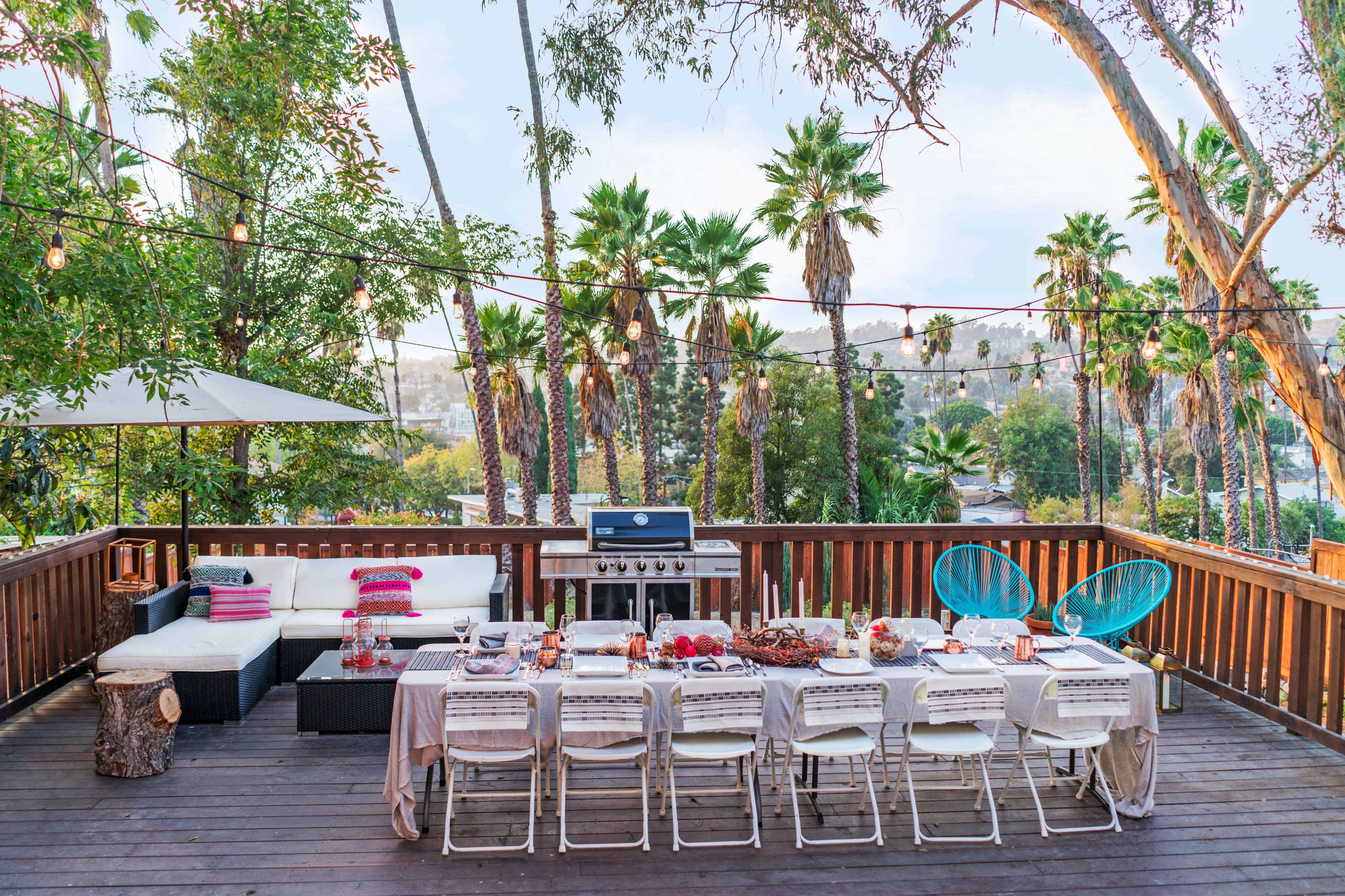 A deck is set for an outdoor dinner with a long table, chairs, a grill, and palm trees in the background.