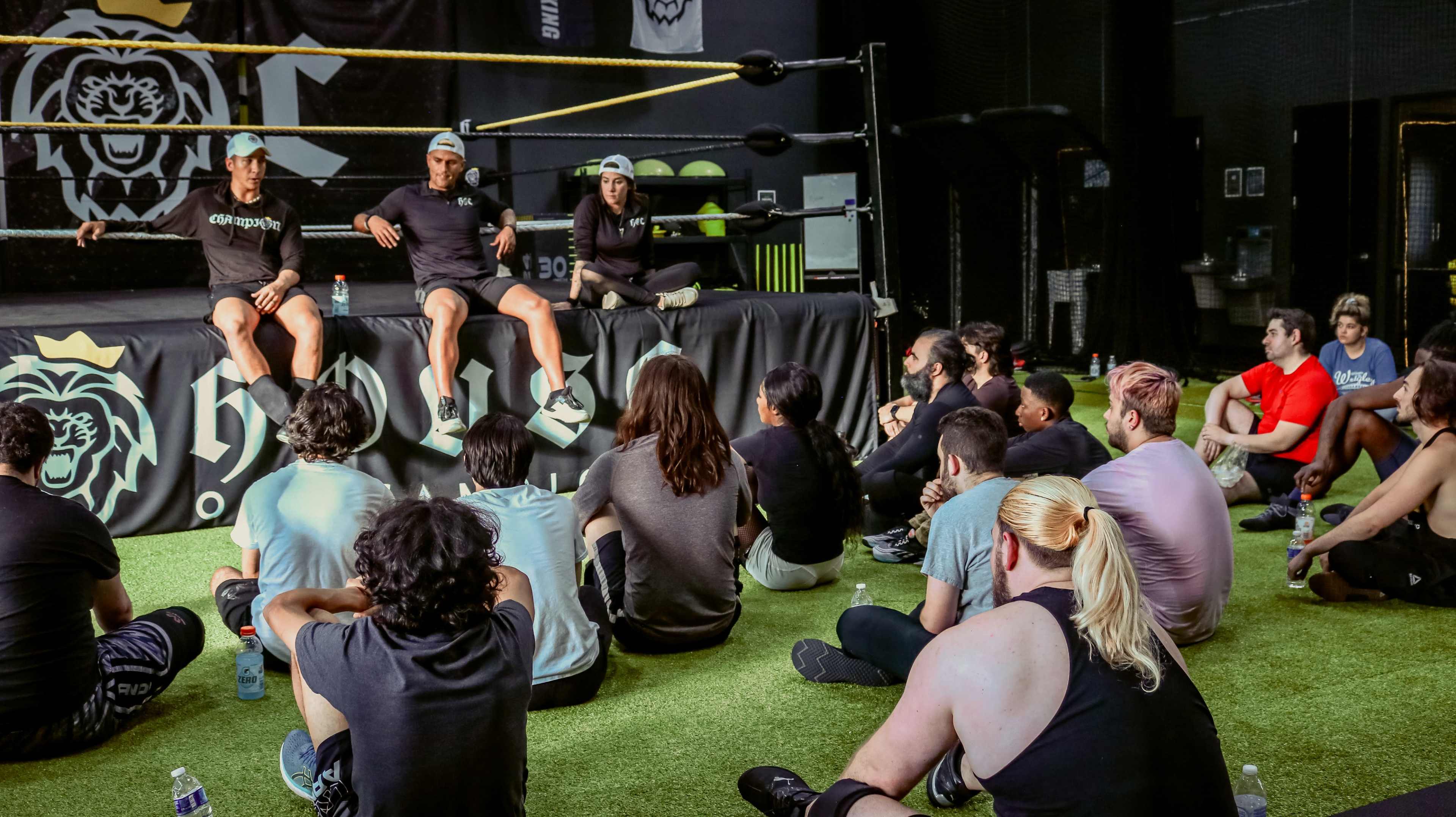 A group of individuals sits on a gym floor, attentively listening to three speakers seated on a wrestling ring.