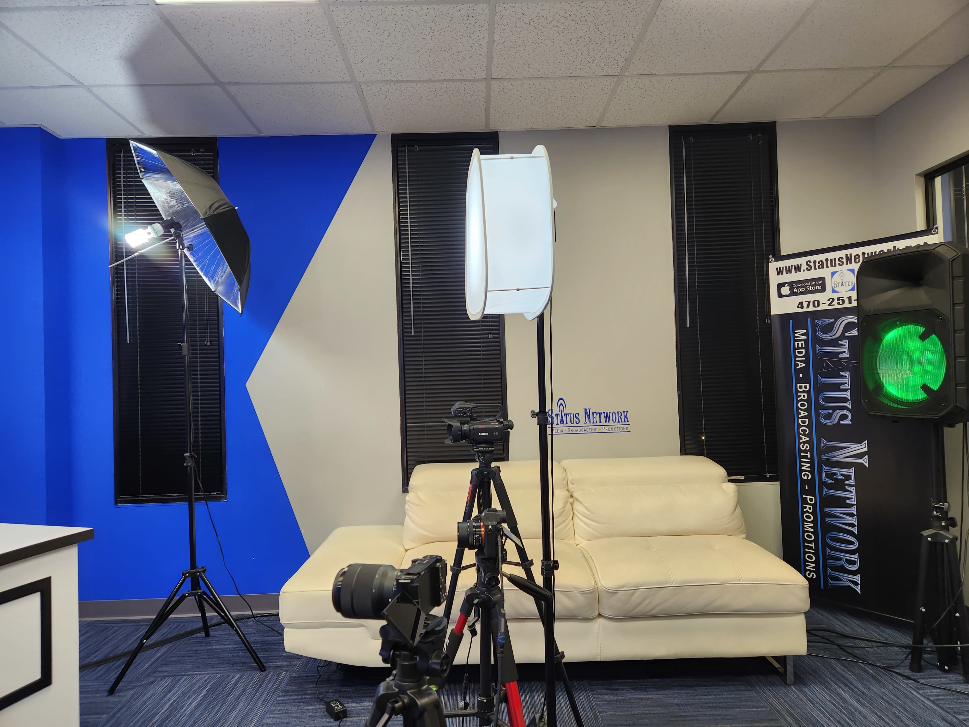 The image shows a studio setup with a white couch, two light sources including a softbox and a ring light, and a camera mounted on a tripod, all against a blue and white wall.