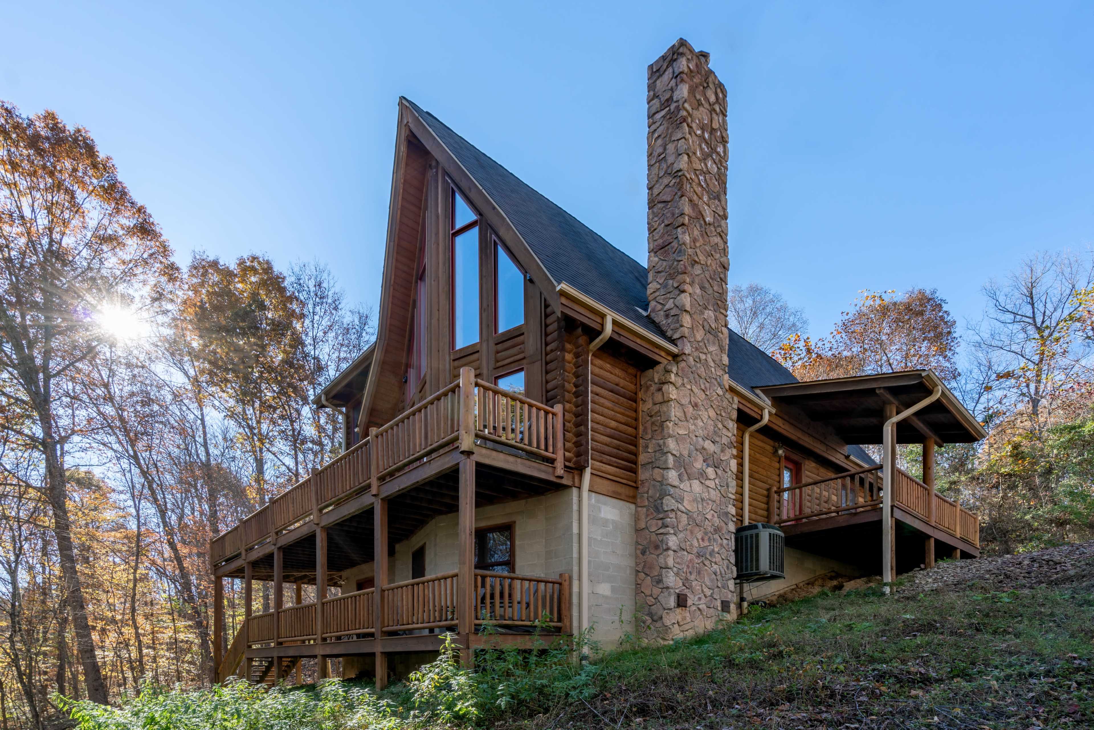 A log cabin with a stone chimney and multiple balconies is situated amid a forested hillside.