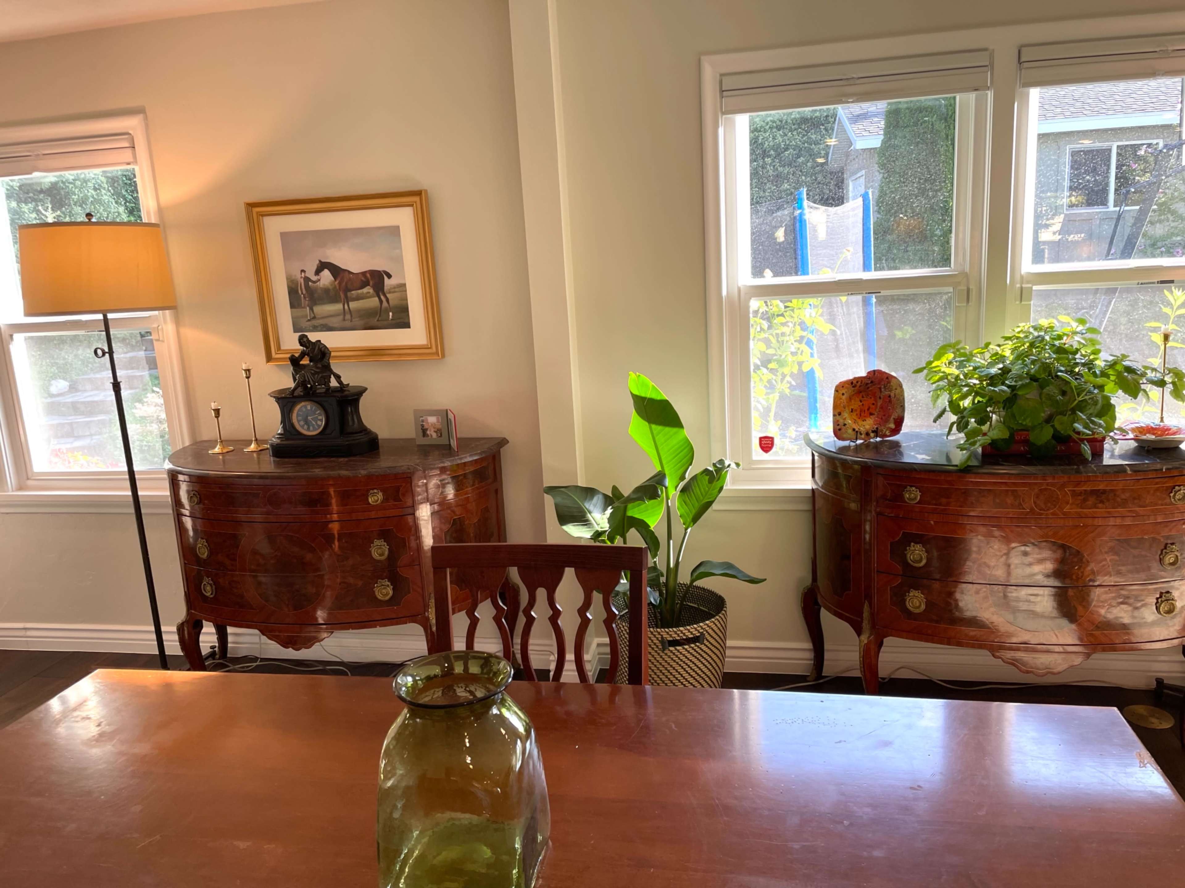 A well-lit room with two ornate side tables, a lamp, a painting of a horse, and a large plant beside a wooden dining table.