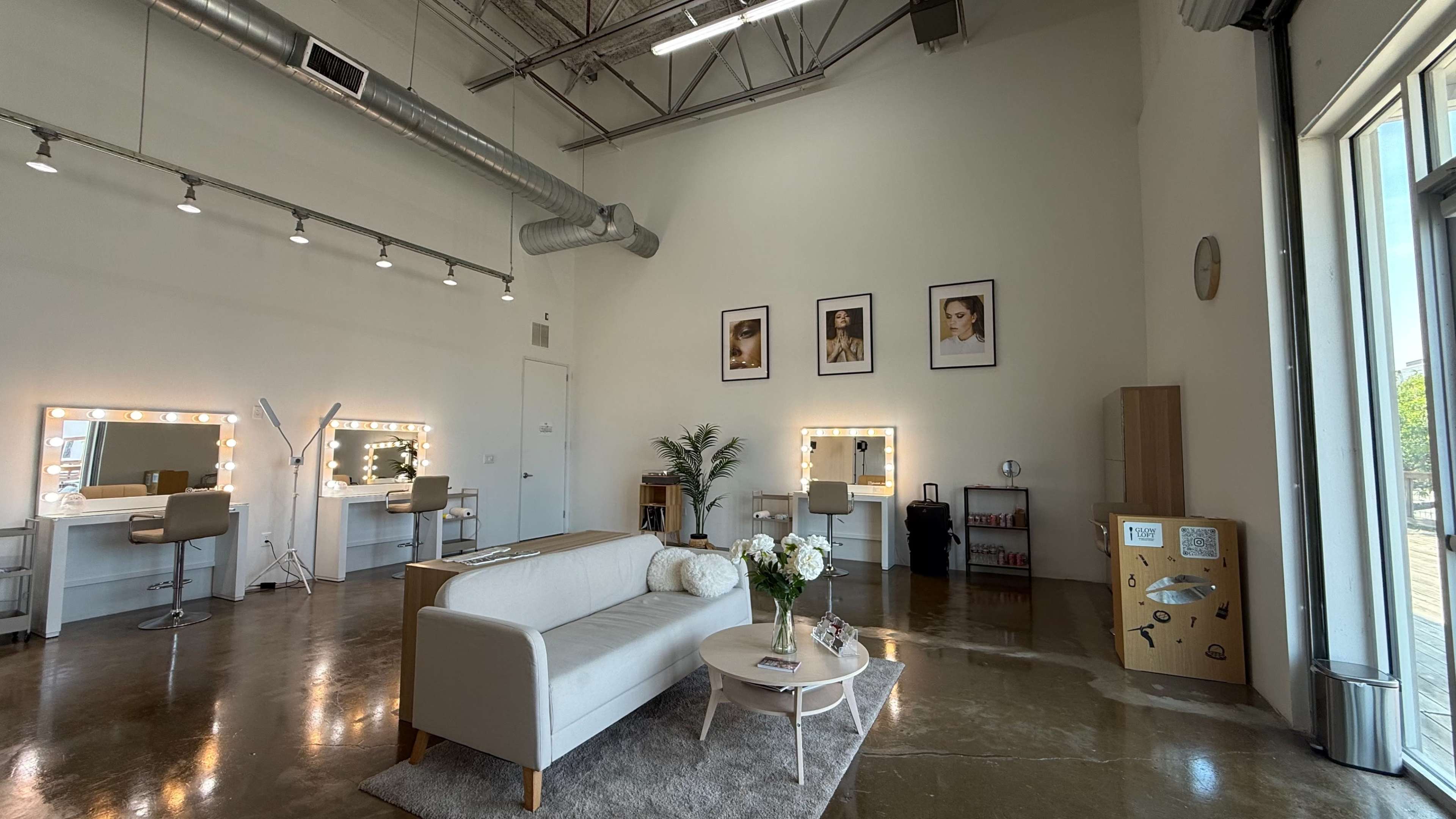 The image shows a modern salon interior with a couch, makeup stations illuminated by lights, and framed photographs on the wall.