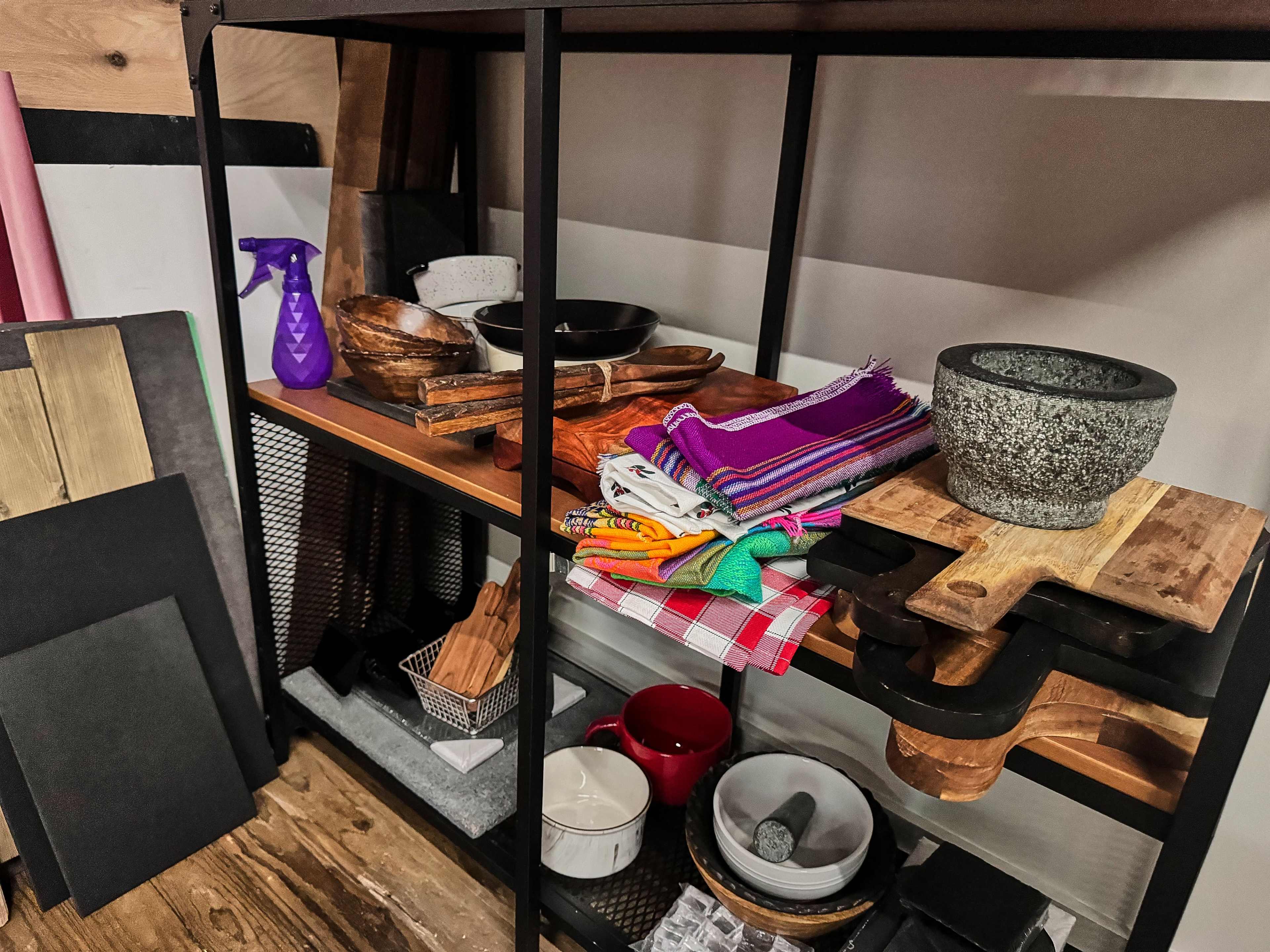 A shelving unit displays a variety of kitchen items, including bowls, cutting boards, colorful textiles, and cleaning supplies.