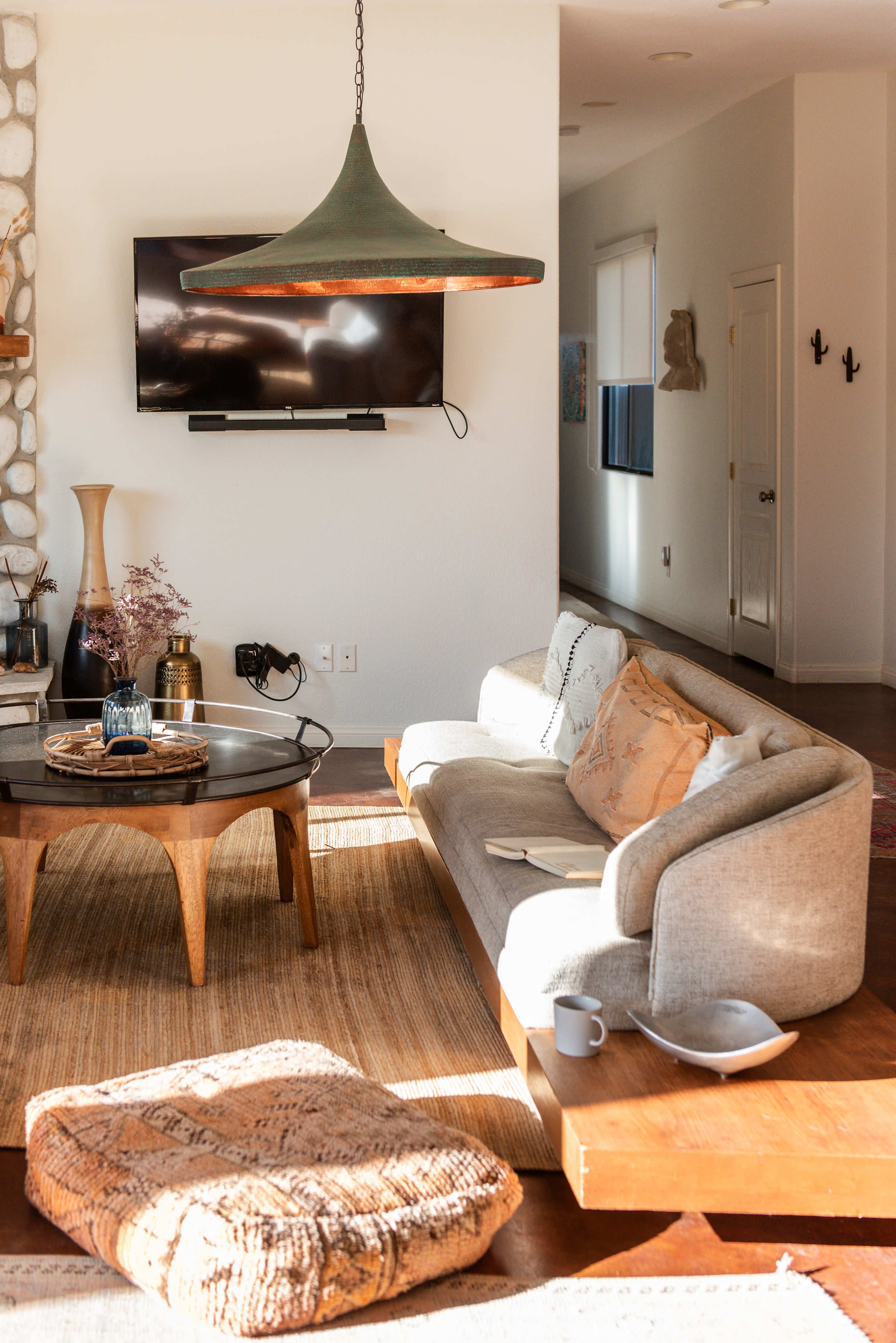 A modern living room features a light gray sofa, a round wooden coffee table, a large pendant light, and a wall-mounted television, with decorative items and a cozy atmosphere.
