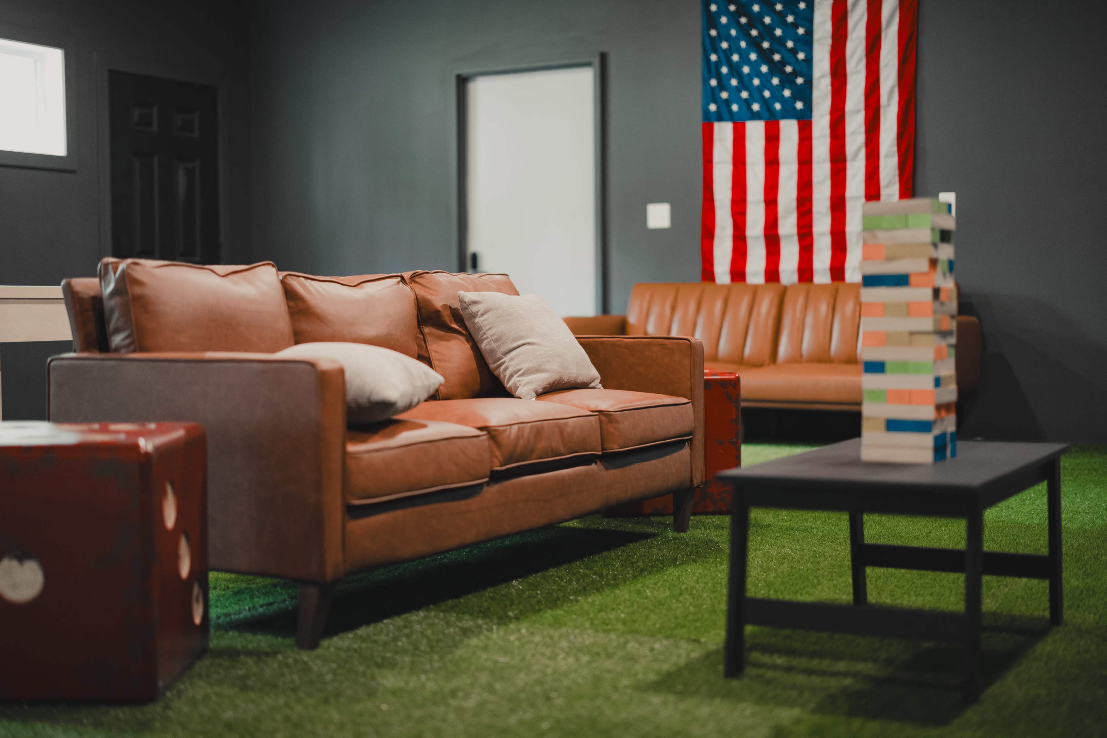 The scene features a cozy seating area with a brown leather sofa, a matching couch, a black coffee table, and a stack of colorful wooden blocks against a backdrop of a US flag on a gray wall.
