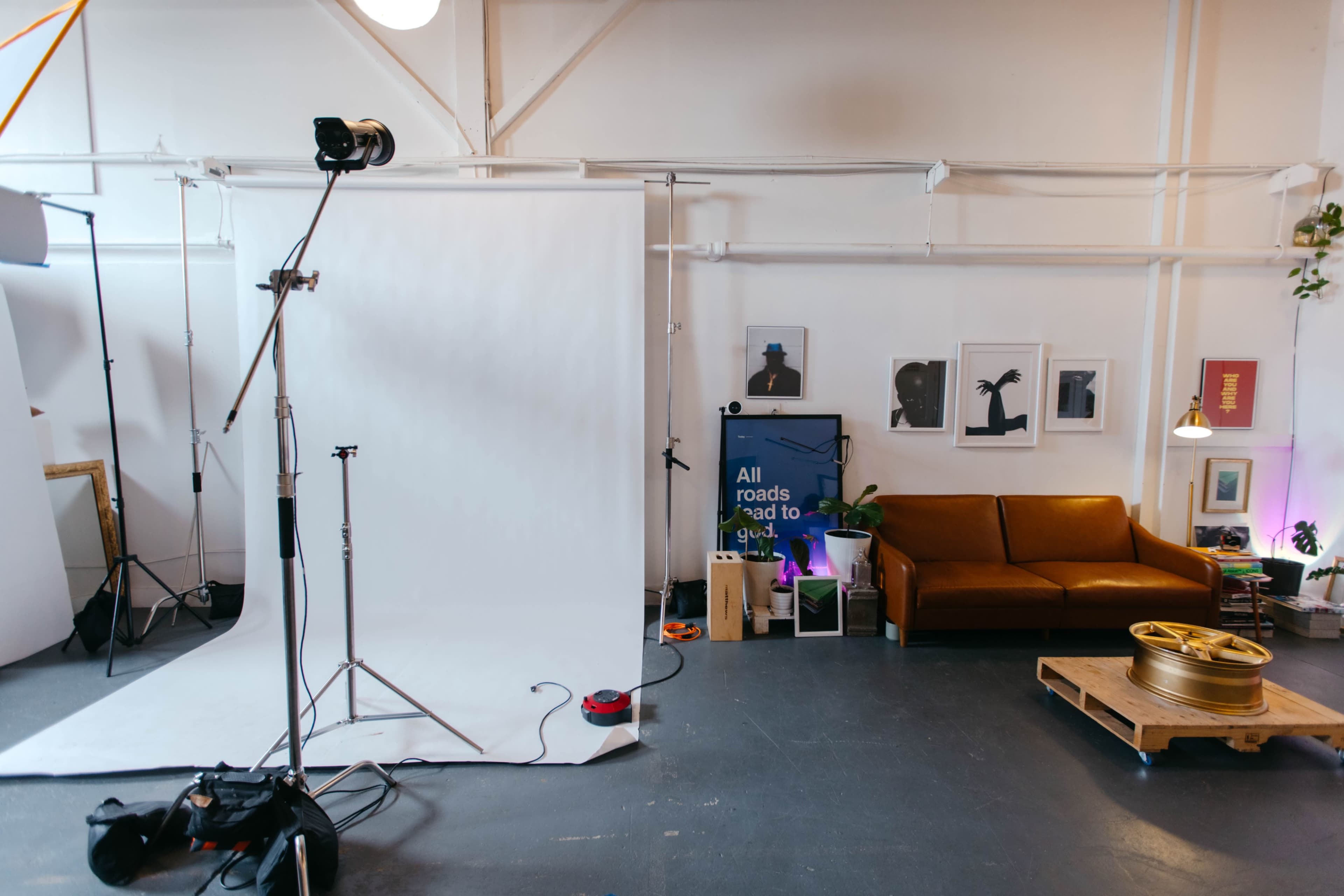 The image shows a studio space featuring a white backdrop, a light stand, a brown couch, and various artworks on the walls.